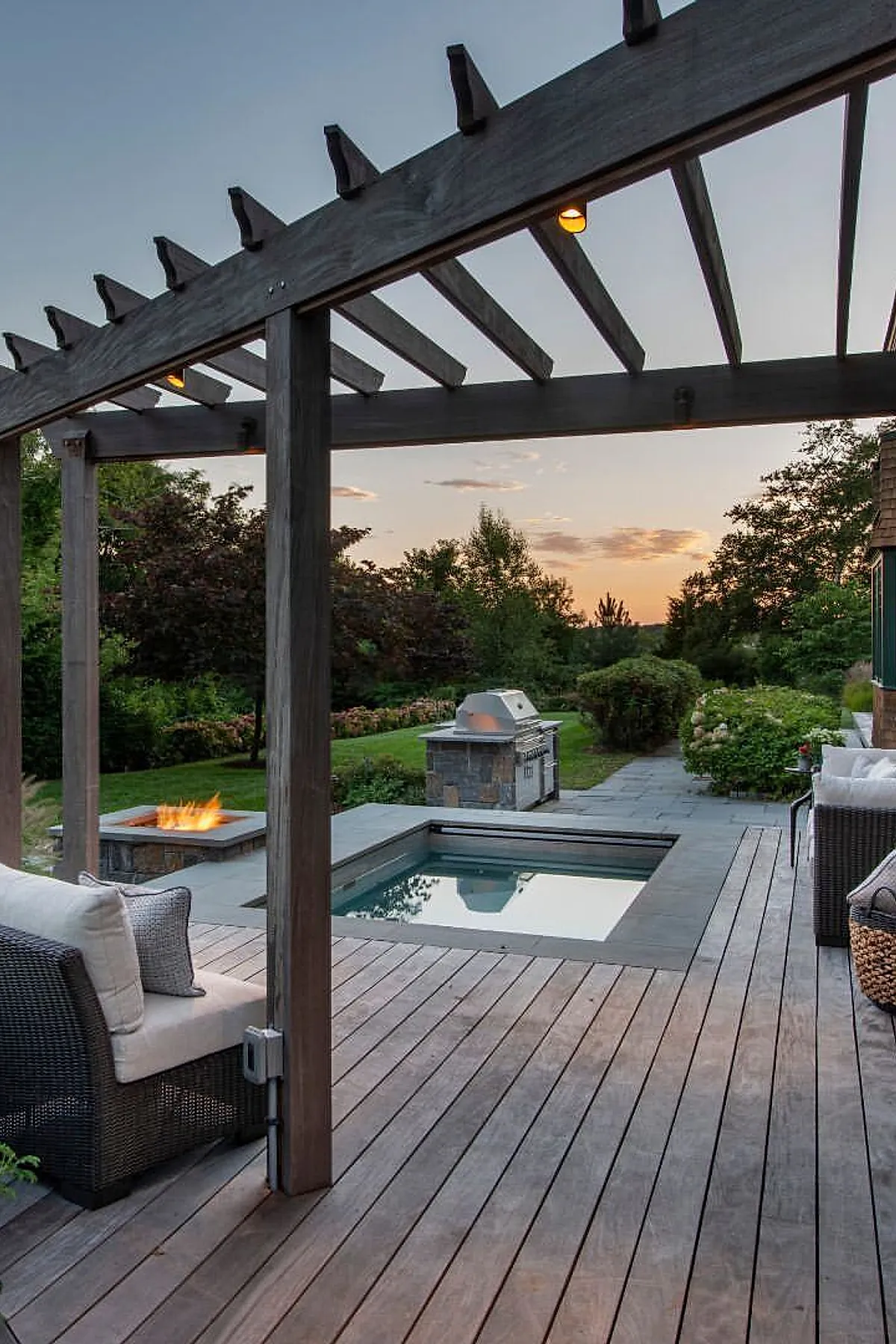 A small inground pool surrounded by stone tiles and wooden decking, featuring a dark wood pergola overhead with built-in lighting, a firepit with flames on one side, an outdoor grill station in the background, and cushioned wicker chairs around the deck with lush greenery beyond.