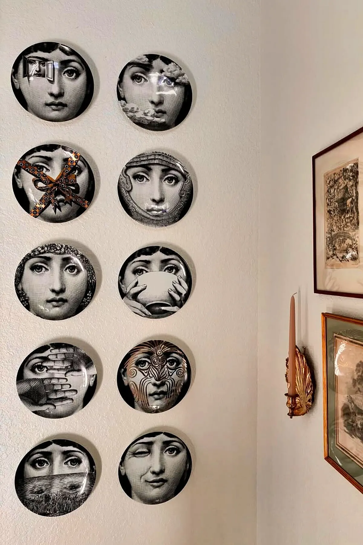 Aesthetic wall decor ideas featuring ten black and white decorative plates with surreal and expressive female faces arranged vertically on a white wall next to framed vintage prints and a gold candle holder.