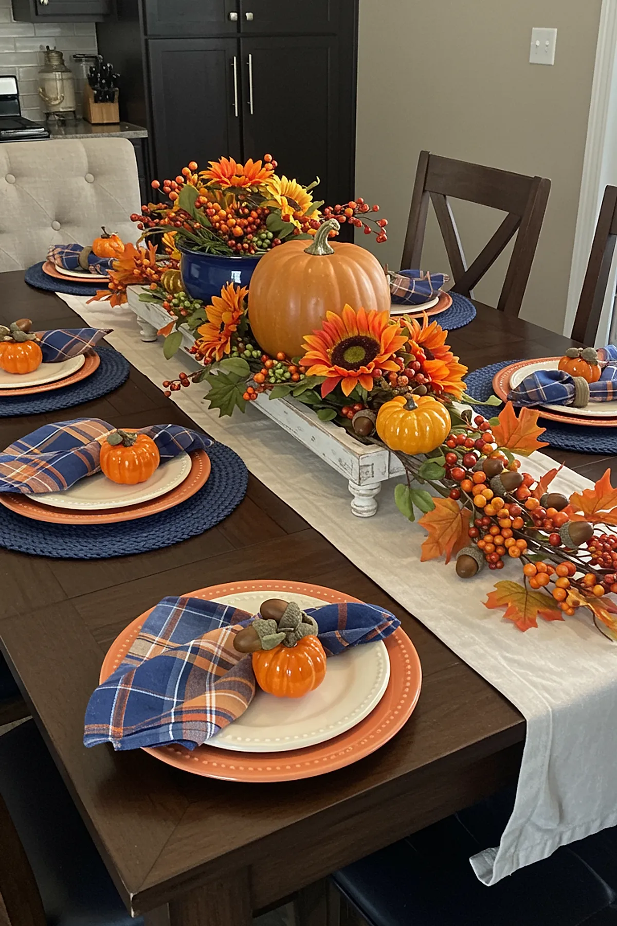 Dark wood dining table set with orange chargers, white plates, blue and orange plaid napkins folded on top, each adorned with a small orange pumpkin with acorn decoration. Centerpiece features large pumpkins, bright sunflowers, berries, and autumn leaves arranged on a cream fabric runner.