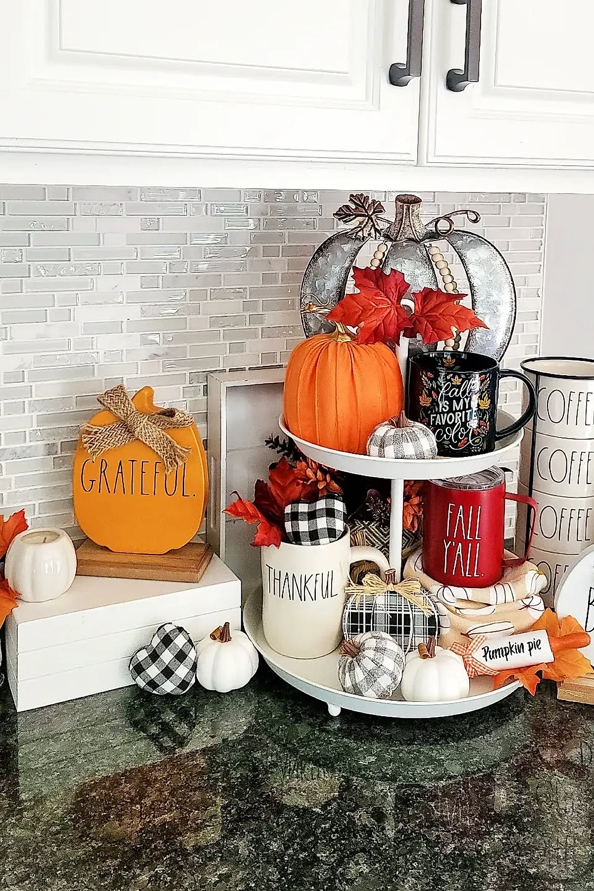 Two-tiered white tray on kitchen counter decorated with orange and white pumpkins, mugs labeled thankful and fall y