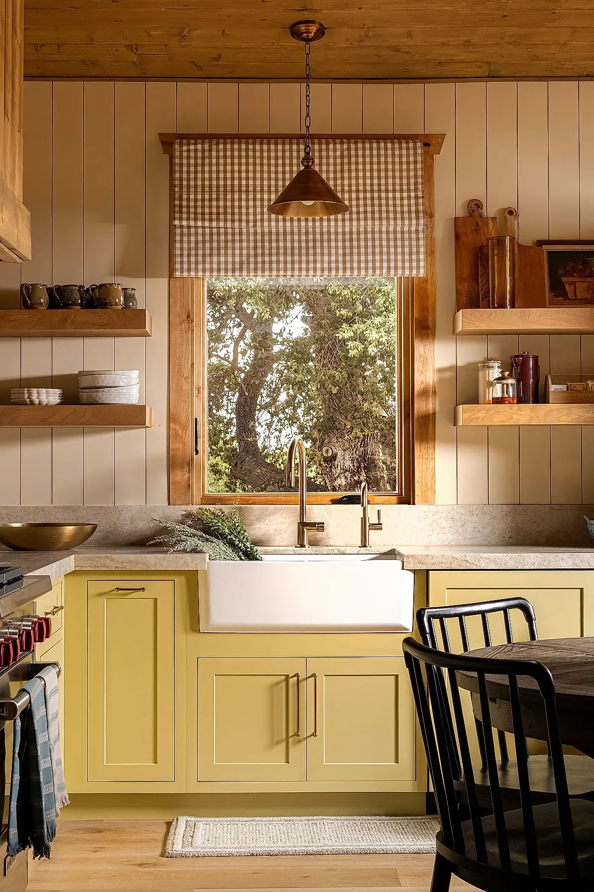 Yellow cottage kitchen featuring light yellow cabinets with brass handles, farmhouse white sink under window with beige gingham curtain, wood shelves with dishes and jars, beige walls and wooden ceiling.
