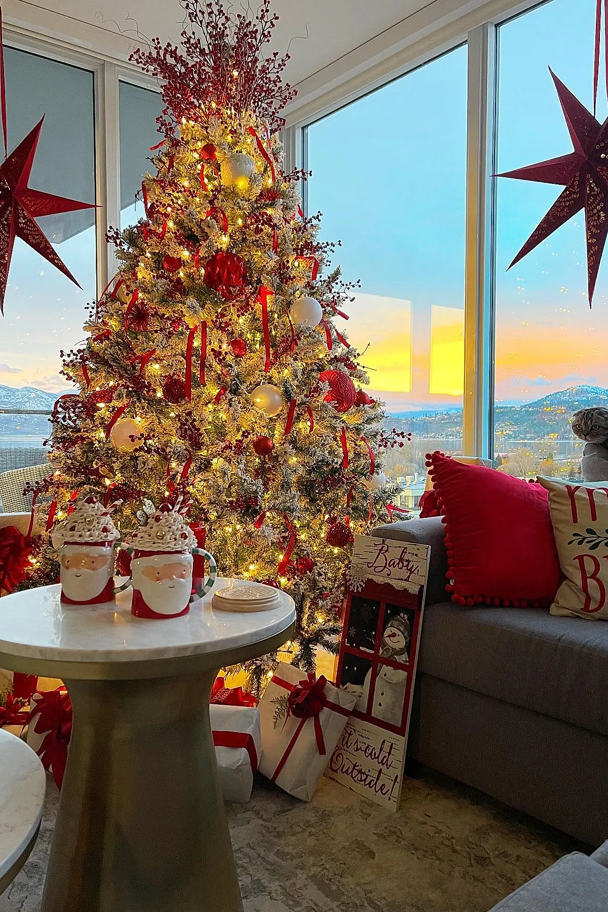 Christmas tree decorated with red ribbons, white and red ornaments, and faux berries beside a gray couch with red pillows and Santa-themed mugs on a round marble table, large glass windows showing sunset background