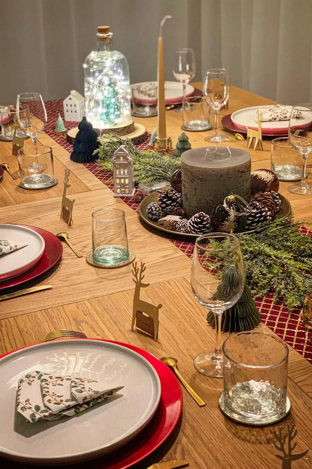 Festively decorated dining table with red tablecloth, floral plates, varied candles, greenery, and fairy lights for a cozy atmosphere.