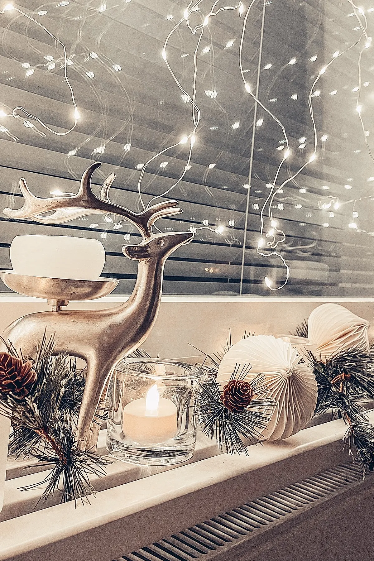 christmas window decorations featuring a metallic reindeer candle holder, a lit tea light candle in a glass holder, pine garland with pine cones, and white paper ornaments on a windowsill with string lights reflected on the window blinds