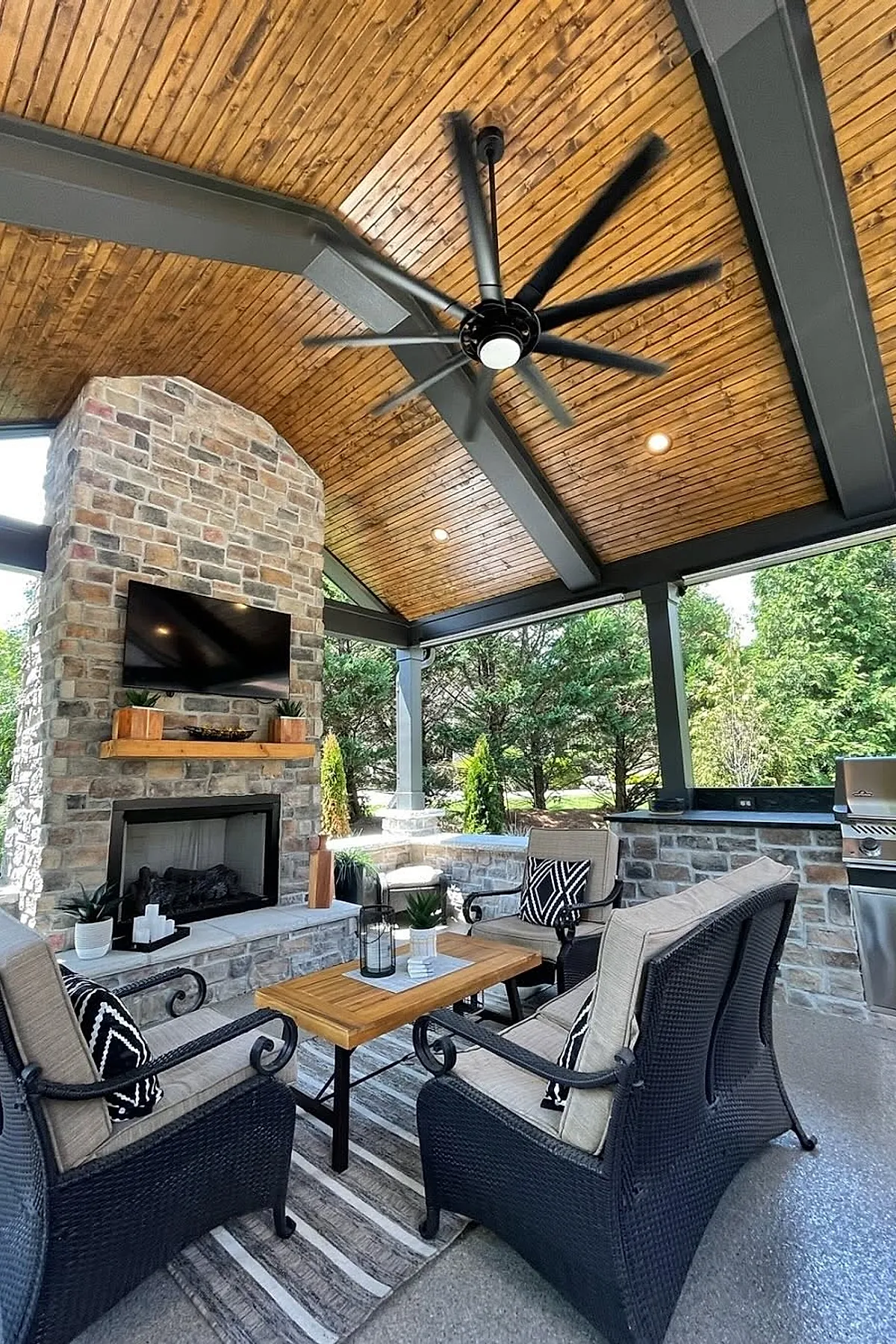 Wooden backyard gazebo with natural wood ceiling and dark beams, featuring a tall stone fireplace with mounted TV above, black wicker cushioned chairs with patterned pillows around a wooden coffee table on striped rug, surrounded by greenery.