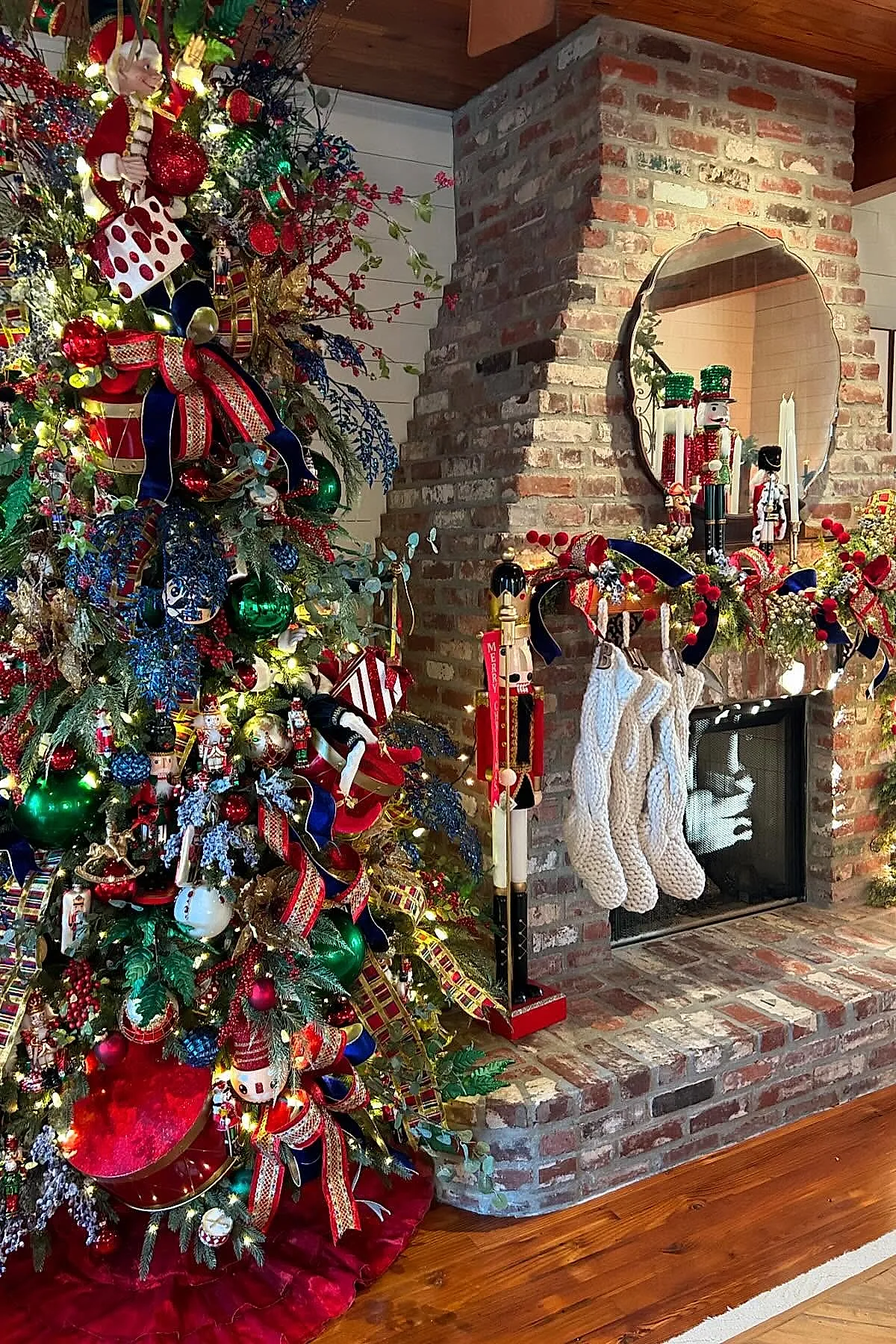 brick fireplace mantle decorated with knitted white Christmas stockings hanging from a lush garland adorned with red berries and navy ribbons, flanked by tall nutcracker figurines, next to a colorful, heavily decorated Christmas tree with ribbons, ornaments, and lights