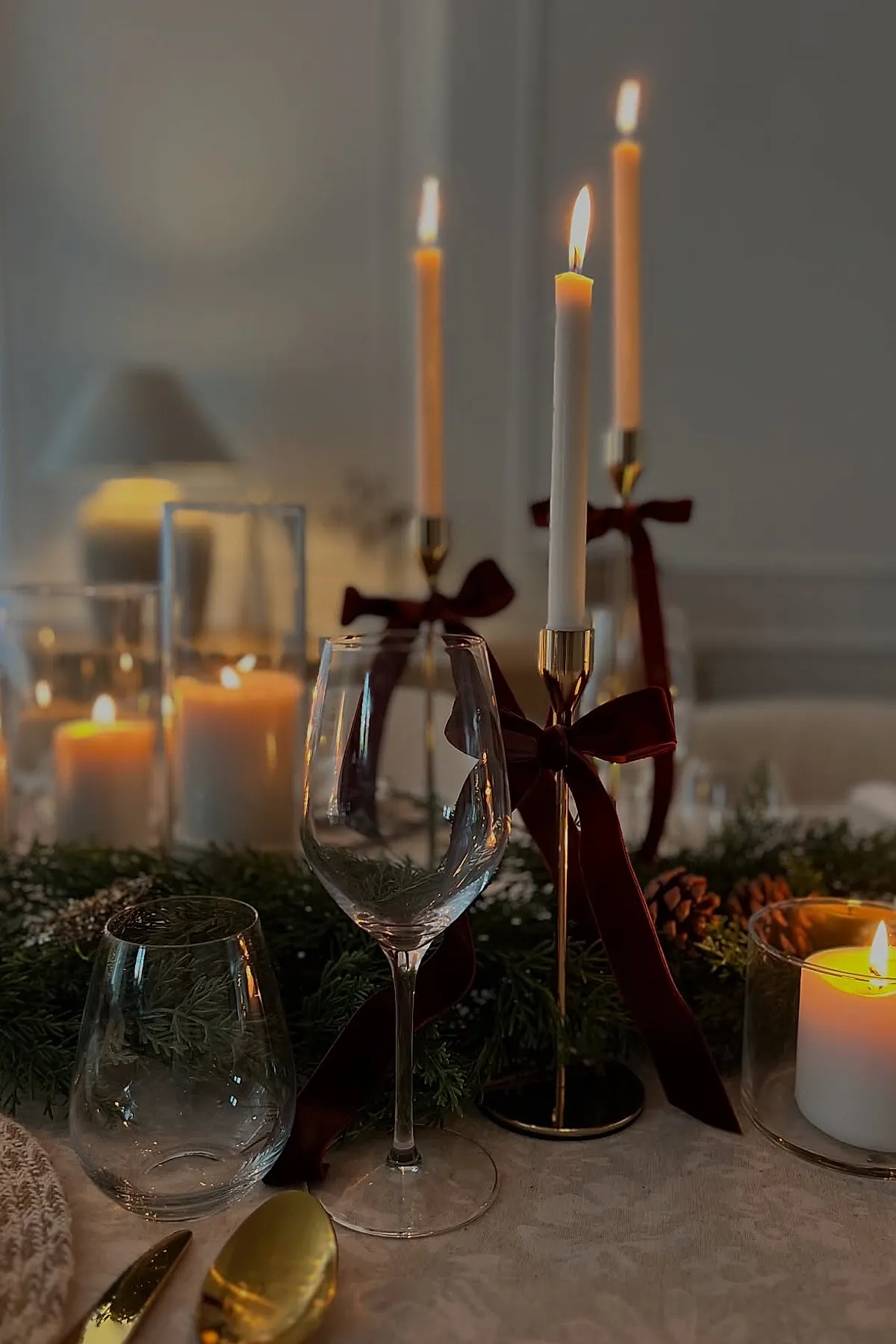 christmas dinner table settings featuring white taper candles in gold candlesticks tied with dark red velvet ribbons, surrounded by greenery, pinecones, clear wine and water glasses, and white pillar candles in glass holders on a light beige tablecloth.