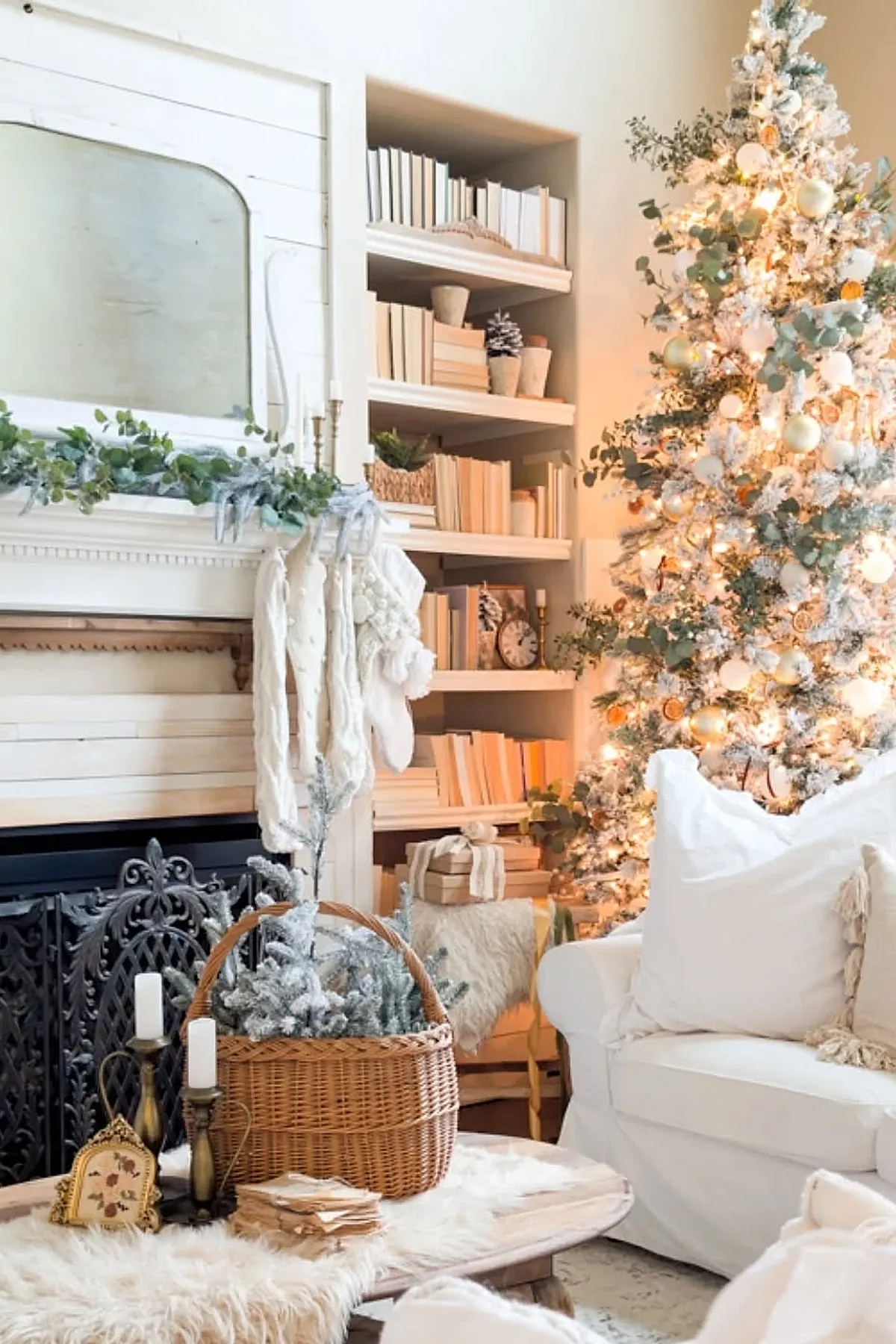 White flocked Christmas tree decorated with white and gold ornaments, eucalyptus sprigs, warm white lights next to white couch, bookshelf filled with neutral books, fireplace mantel with greenery and hanging cream knit stockings.