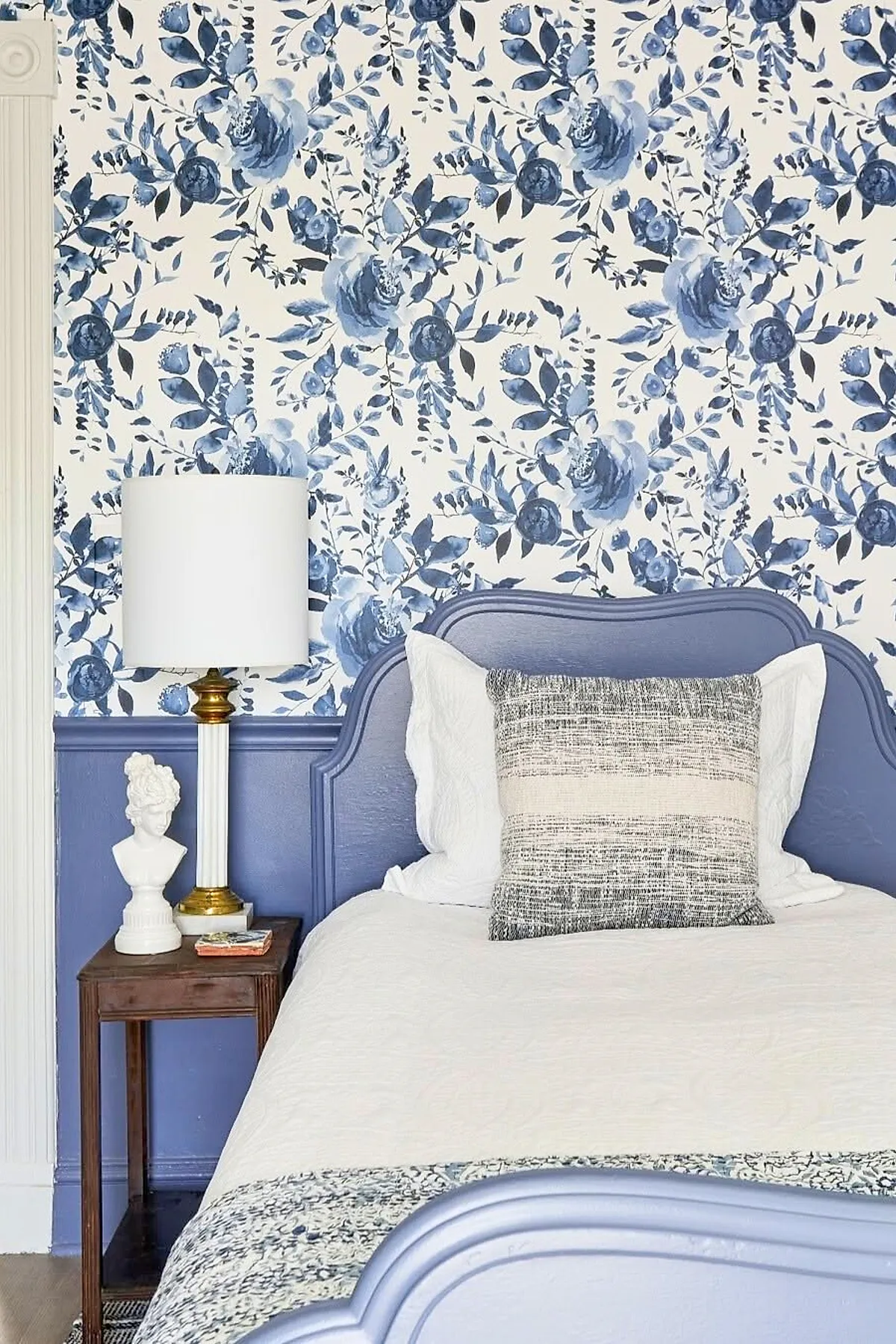 French Blue bedroom featuring floral patterned wallpaper in shades of blue and white, a French blue wooden bed frame with curved edges, white bedding with textured pillows, a dark wood vintage nightstand holding a white lamp with gold accents and a small bust sculpture.
