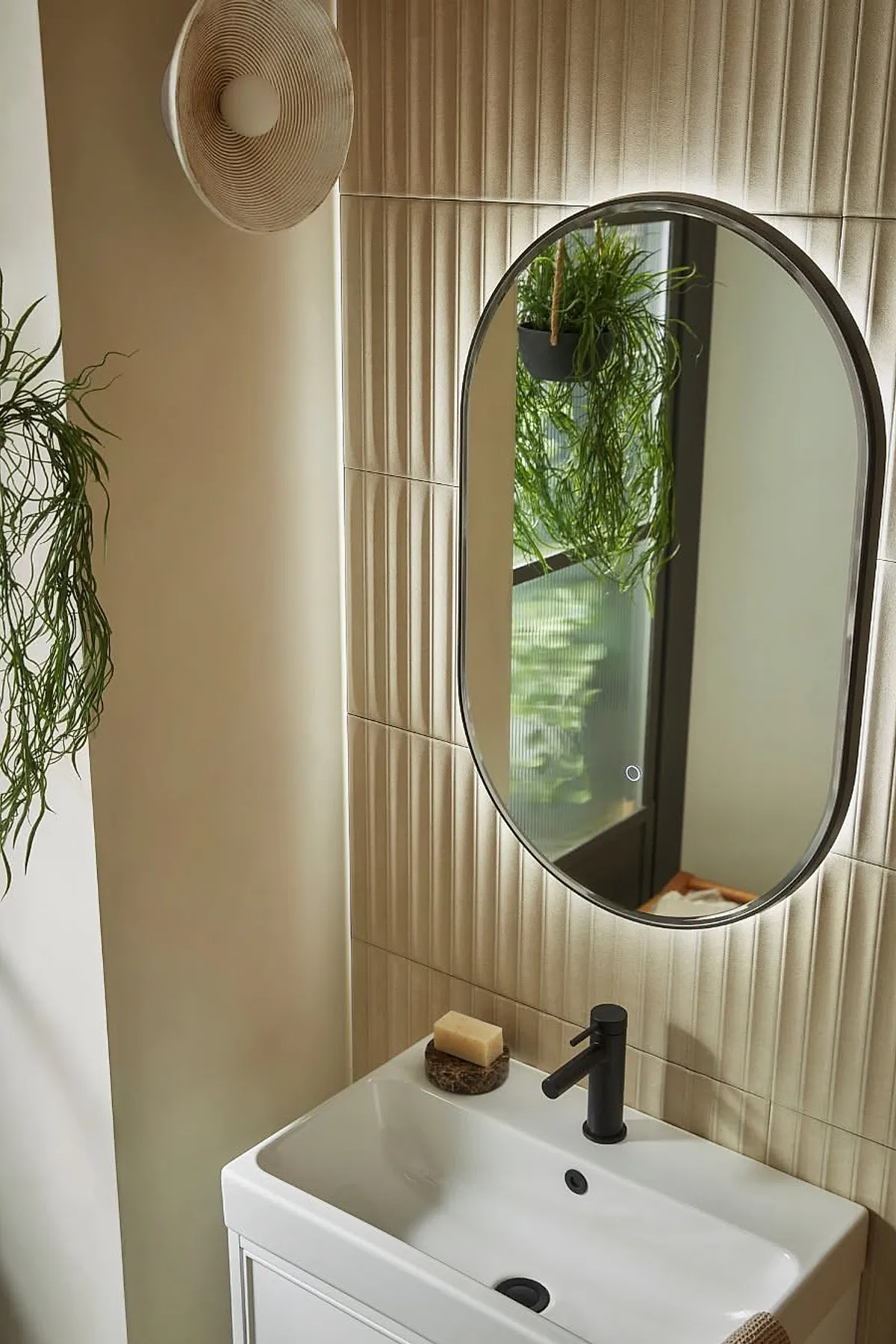 Modern bathroom with an oval backlit mirror mounted on vertically ridged beige tiles, featuring a white sink with a black faucet and green hanging plant reflected in the mirror.