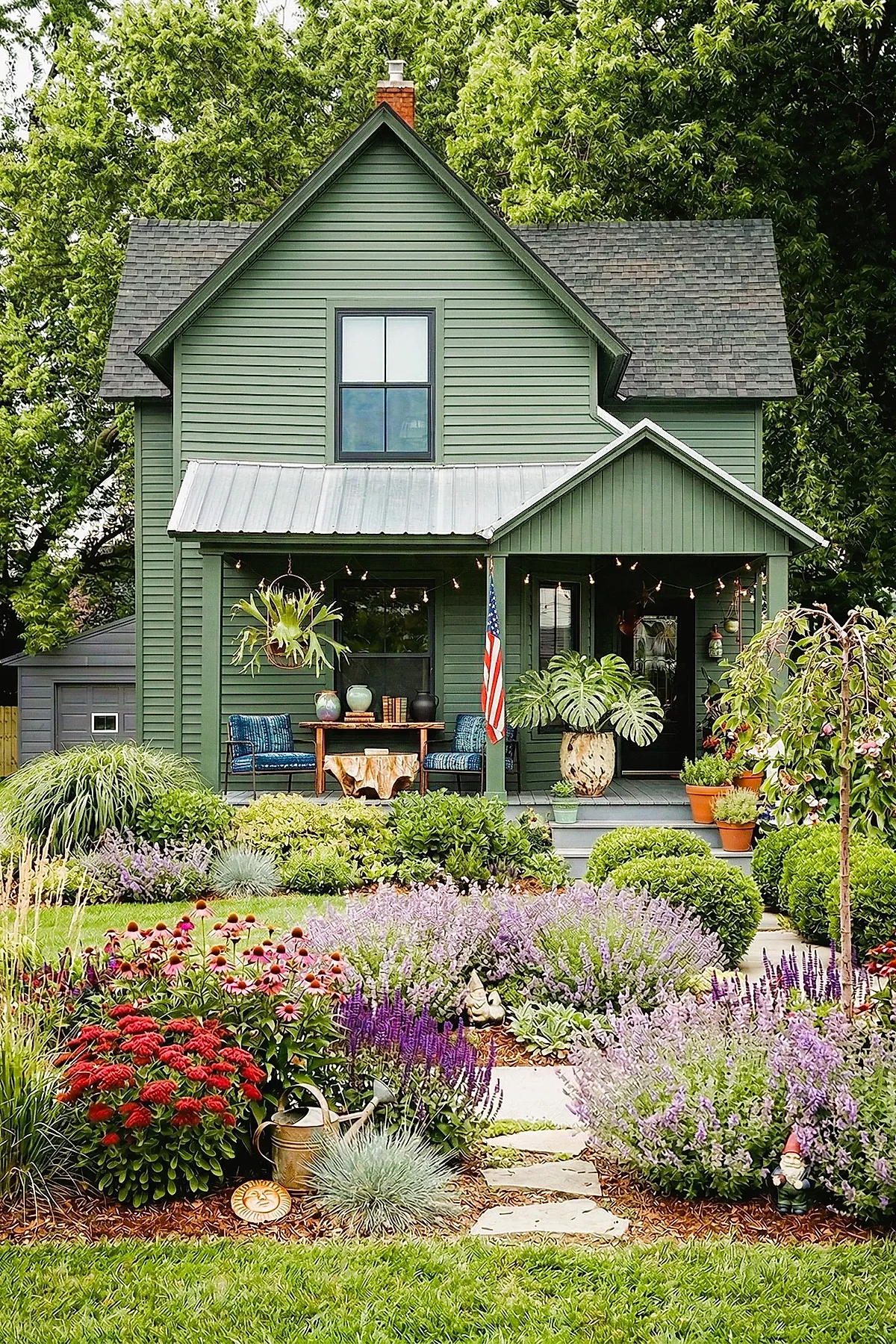 Small front yard landscaping featuring vibrant flower beds with purple, red, and pink blooms arranged along a stone pathway leading to a green house with porch seating and potted plants.