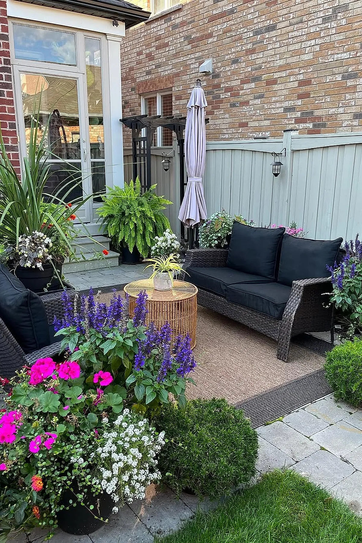 Small patio with black rattan sofa featuring black cushions, surrounded by vibrant purple and pink flowering plants, a round wicker coffee table with a small green plant on top, beige outdoor rug, light gray wooden fence and brick wall background.