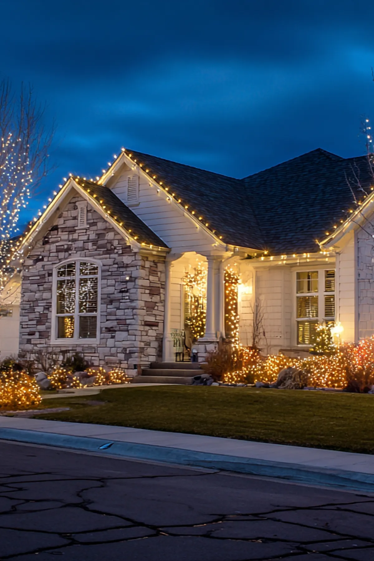 Stone and white siding house exterior decorated with warm yellow outdoor christmas lights outlining roof peaks, porch columns wrapped in lights, illuminated bushes, and trees glowing against a deep blue twilight sky.
