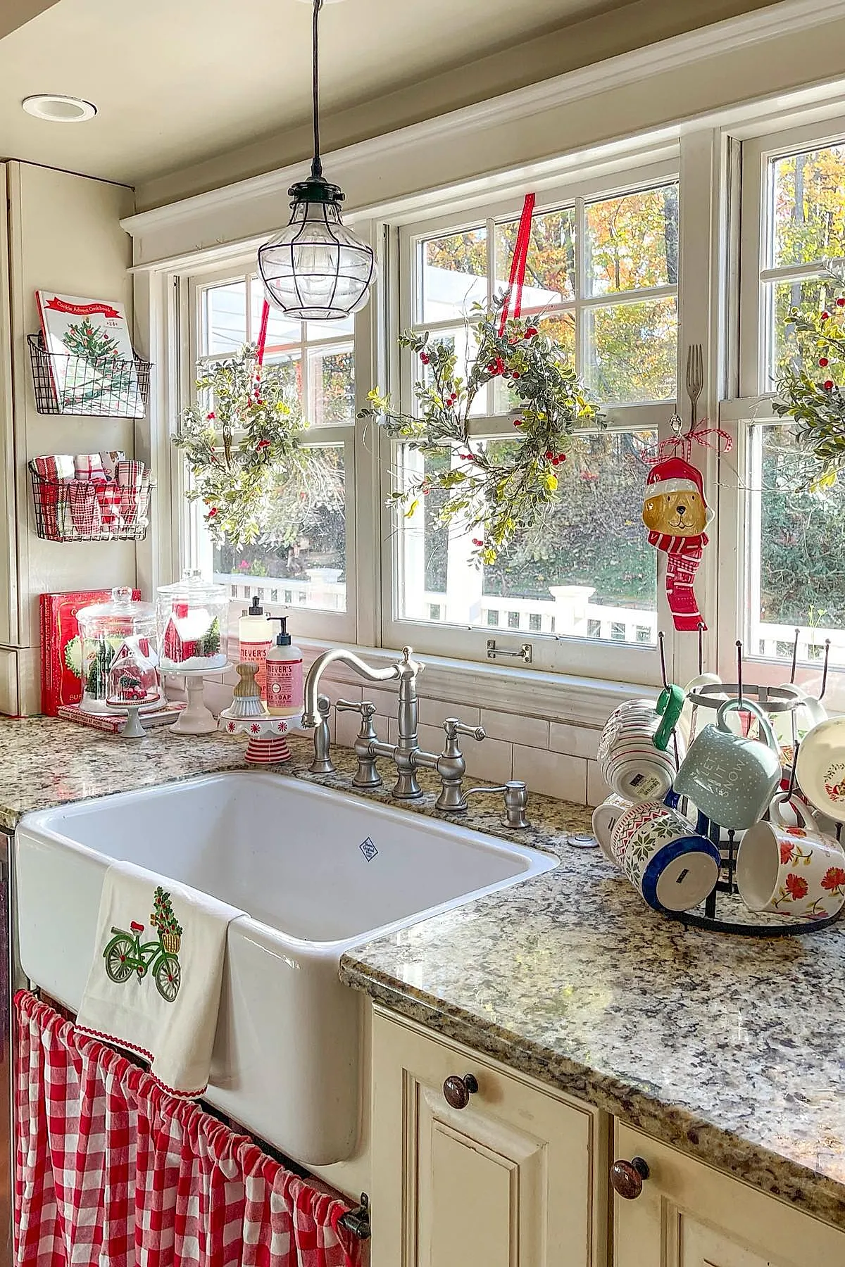 christmas window decorations featuring three green wreaths with red berries hanging from red ribbons on white framed windows above a granite countertop kitchen sink, surrounded by holiday-themed towels, dish soap, glass domes with miniature holiday scenes, and colorful mugs drying on a rack