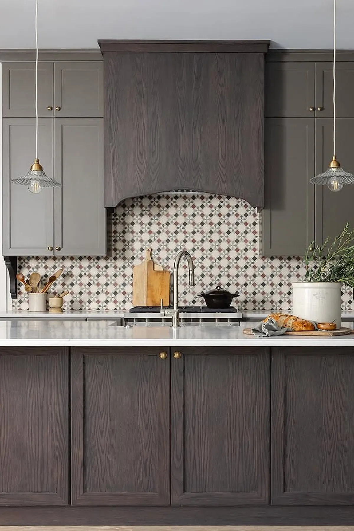 Kitchen with dark wood island cabinets and light gray upper cabinets, decorated with a cutting board holding bread, a white ceramic pot filled with green foliage on a white countertop, set against patterned tile backsplash and illuminated by two hanging pendant lights.