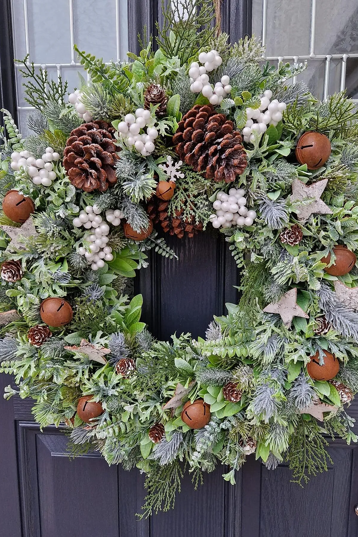 Christmas wreath made from mixed frosted evergreen branches, large brown pinecones, clusters of white berries, rusty jingle bells, and pale wooden star ornaments hung on a dark door.