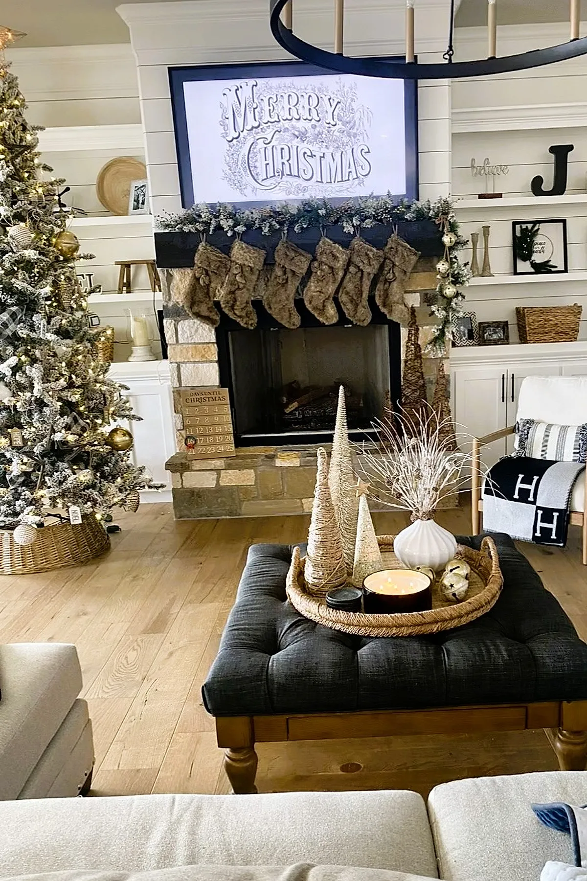 christmas living room with stone fireplace decorated with garland of frosted greenery, six fuzzy brown stockings hanging from the mantel, a large flocked Christmas tree with white and gold ornaments in woven basket stand, light wood flooring, and a dark tufted ottoman holding natural decorative trees and candle tray