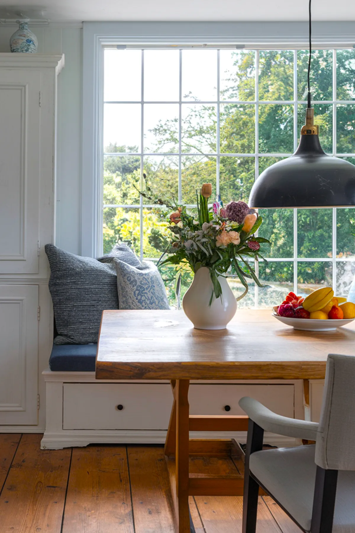 Dining table featuring a floral arrangement and a bowl of fresh fruits by a sunny window.