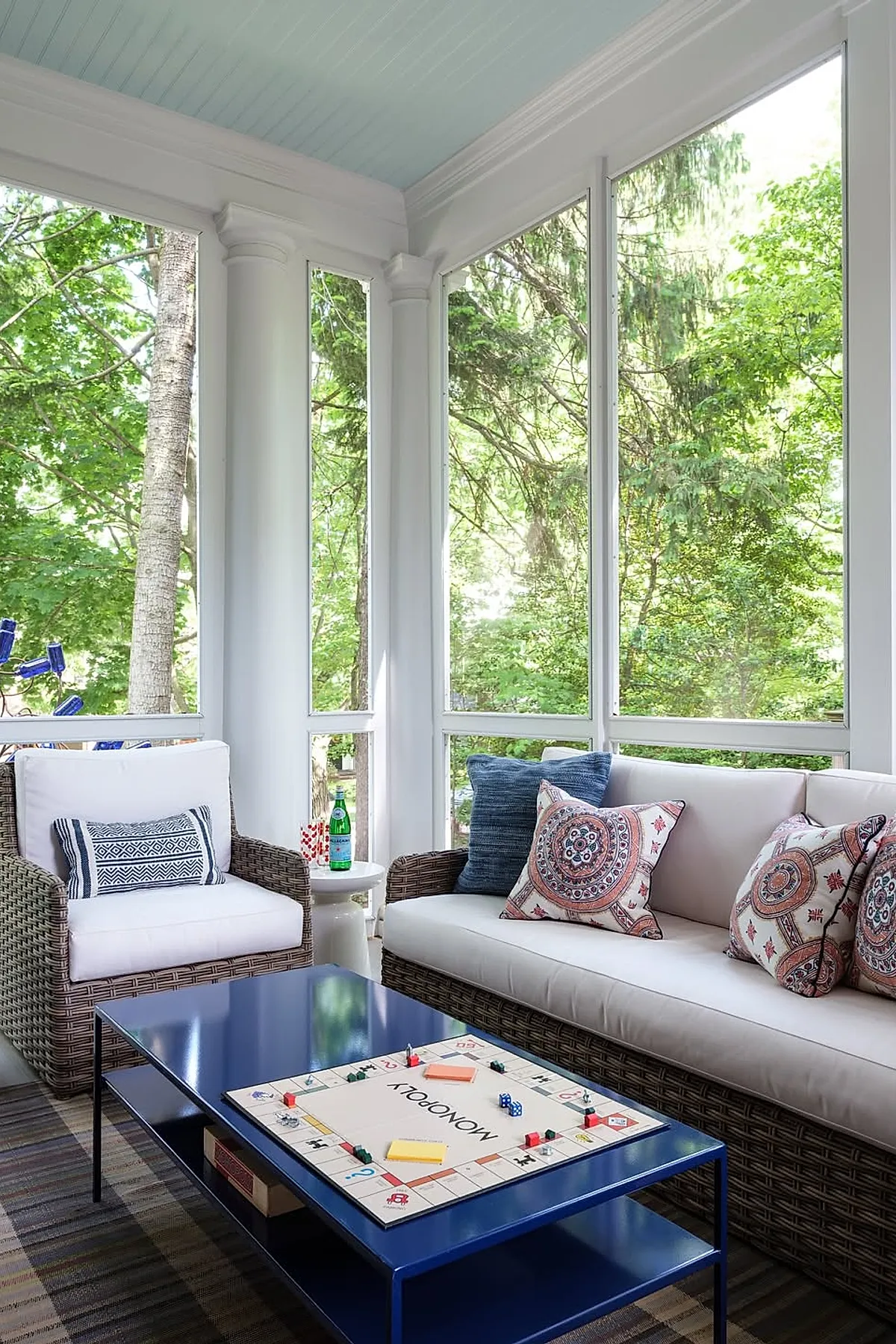Screened in porch featuring wicker armchair and sofa with white cushions and patterned pillows, blue coffee table with Monopoly board game, surrounded by large screened windows overlooking green trees