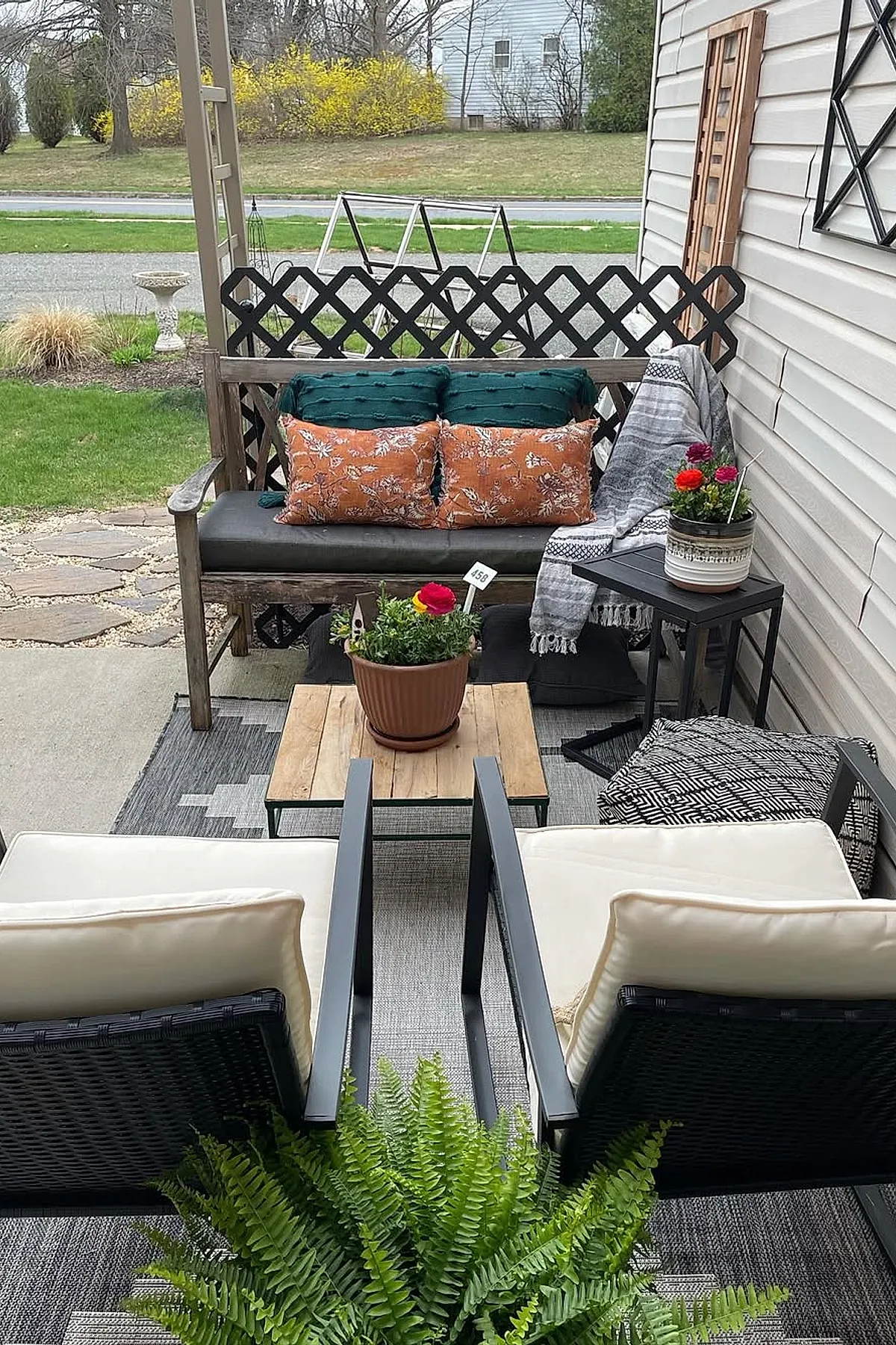 Small outdoor patio with a wooden bench adorned with green and orange patterned cushions, two cushioned black chairs facing a wooden coffee table with potted plants, black side table holding a colorful flower pot, gray patterned rug, and lattice panels against the house wall surrounded by greenery.