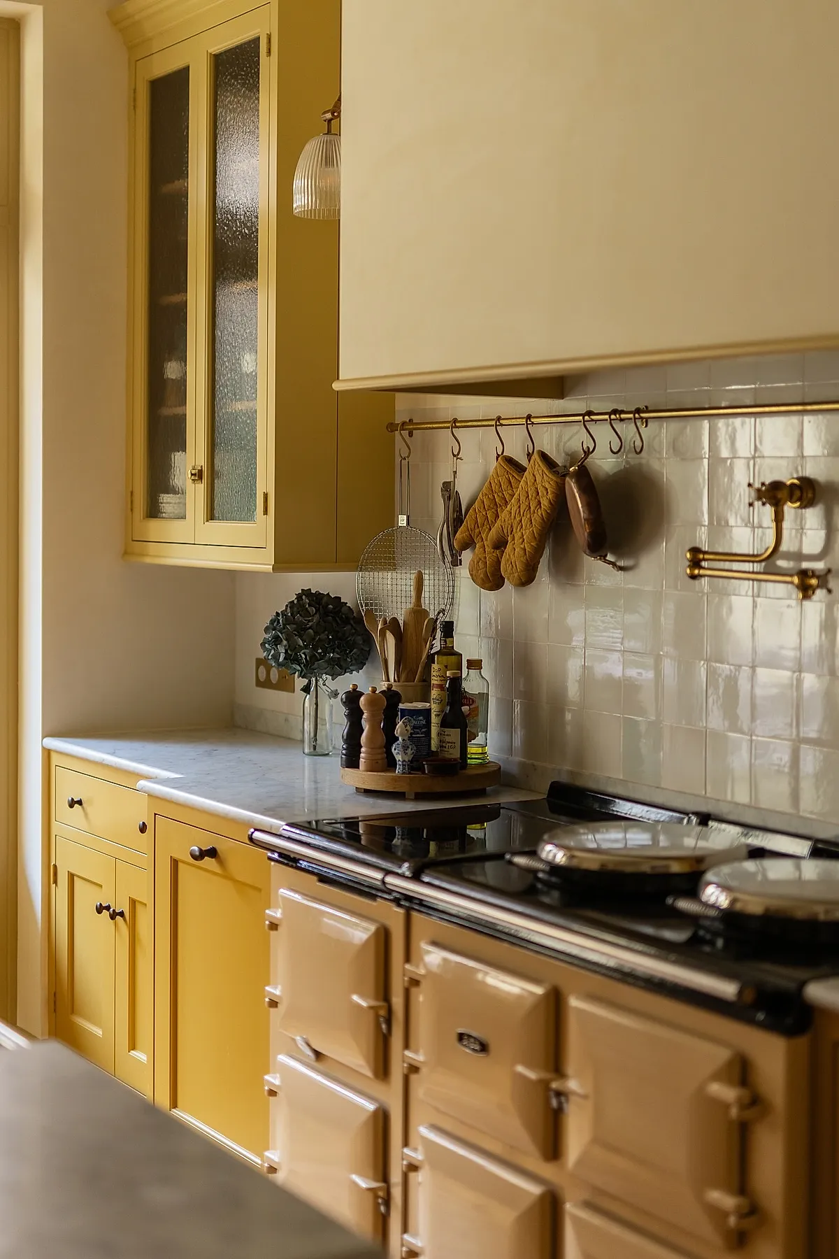 Vintage yellow kitchen featuring light yellow kitchen walls, textured glass cabinet doors, mustard yellow lower cabinetry, cream-colored vintage oven with multiple compartments, brass fixtures, marble countertop holding wooden utensils and condiments, and a hanging rod with mustard oven mitts against a beige tiled backsplash.