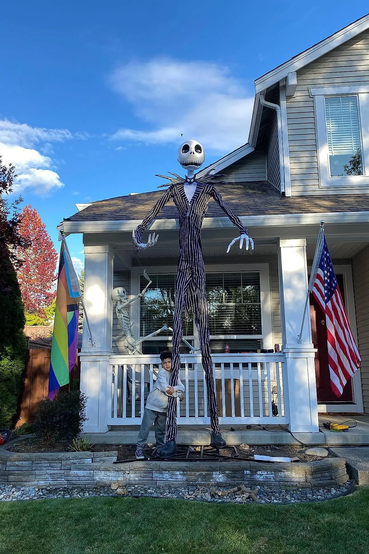 Very tall Jack Skellington figure wearing black and white pinstripe suit standing in front yard porch next to child hugging its leg, with skeleton prop sitting on porch railing, rainbow and American flags on either side.