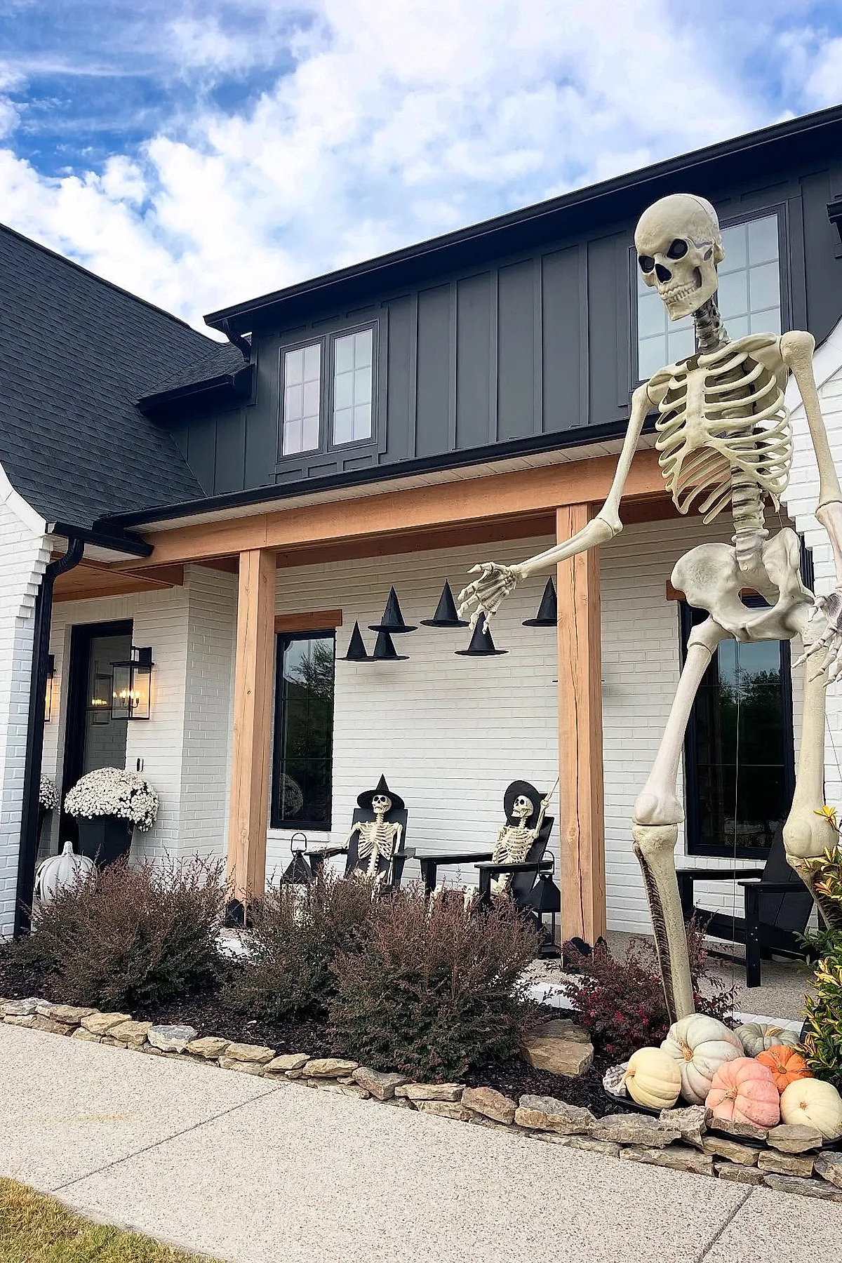 Large standing skeleton next to two seated skeletons wearing witch hats on porch of white brick house with wooden posts, black hanging witch hat decorations overhead, and pale colored pumpkins on stone border.