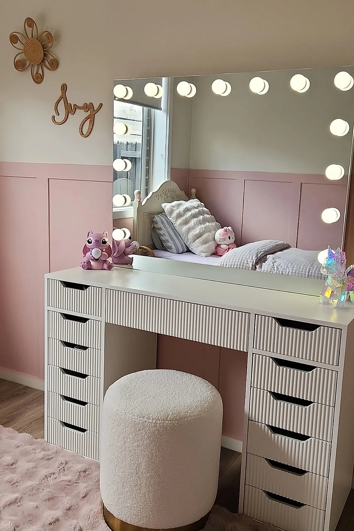 white vanity desk with vertical ribbed drawer fronts, large rectangular mirror framed by round white light bulbs, round plush white stool with gold base, pink wall paneling, small purple and clear unicorn figurines on desk