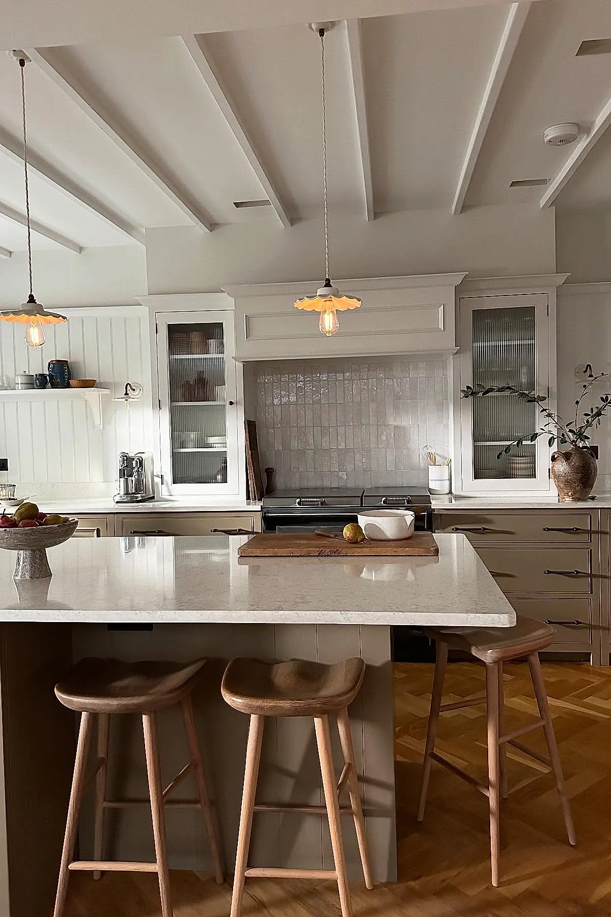 Cottage kitchen featuring glossy white rectangular tile backsplash behind the stove area, cream and taupe cabinetry with glass doors, wooden countertop island with stools, pendant lights with scalloped shades, and a neutral color palette with natural wood flooring.