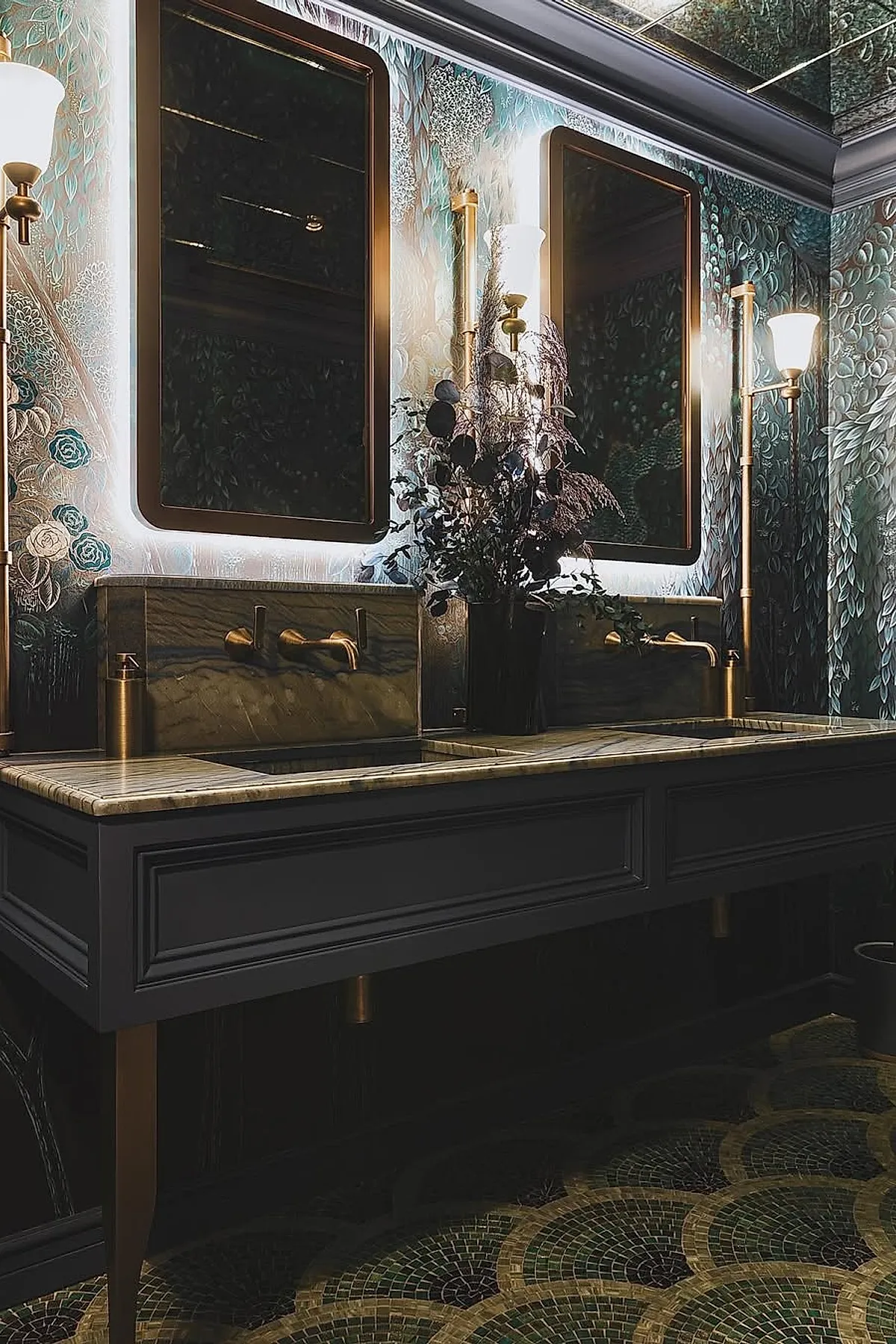 Dark moody bathroom with double backlit rectangular mirrors framed in bronze, brass wall sconces with white shades, a dark wooden vanity with marble countertop, patterned floral wallpaper in teal and silver tones, and a mosaic tiled floor in green and gold hues.