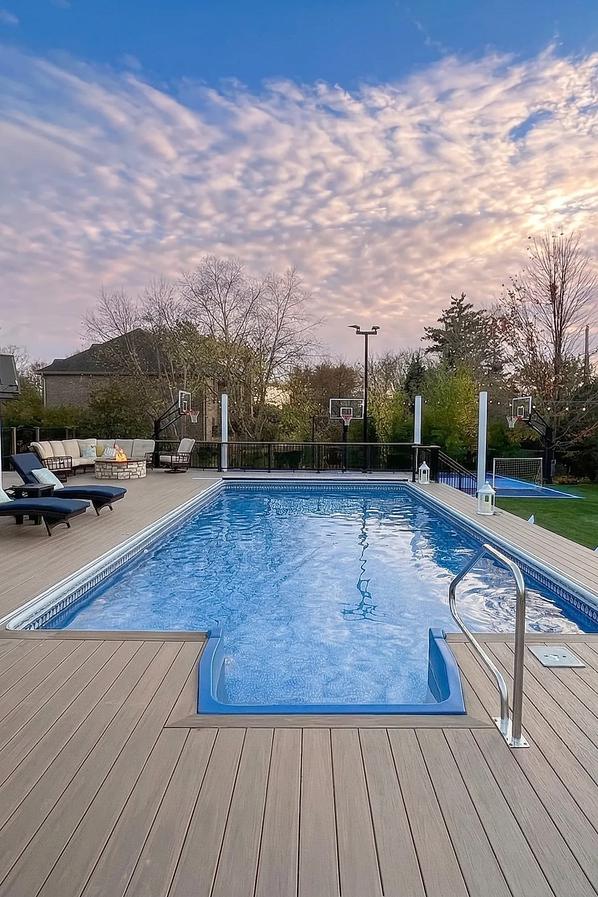 A spacious pool deck with light brown composite decking surrounding a rectangular swimming pool, navy blue cushioned lounge chairs, a stone firepit with beige seating, and multiple basketball hoops in the background under a partly cloudy sky.