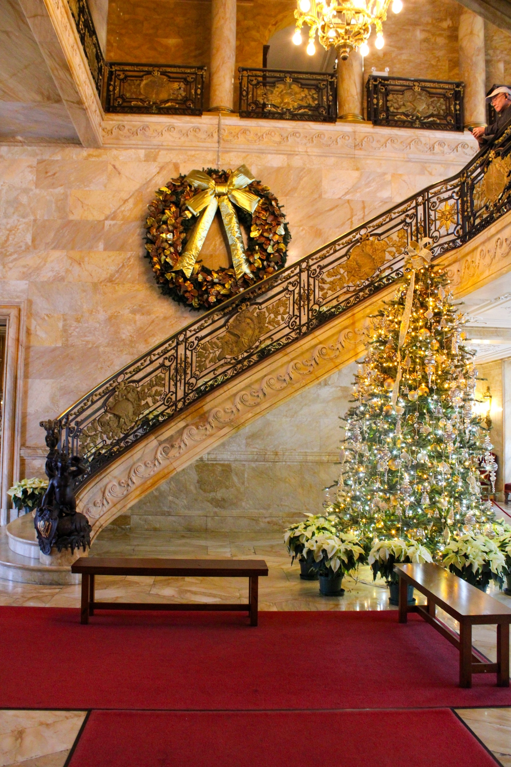 traditional Christmas tree with gold and silver ornaments, white lights, and poinsettias at the base beside a grand marble staircase with gold railing and large wreath