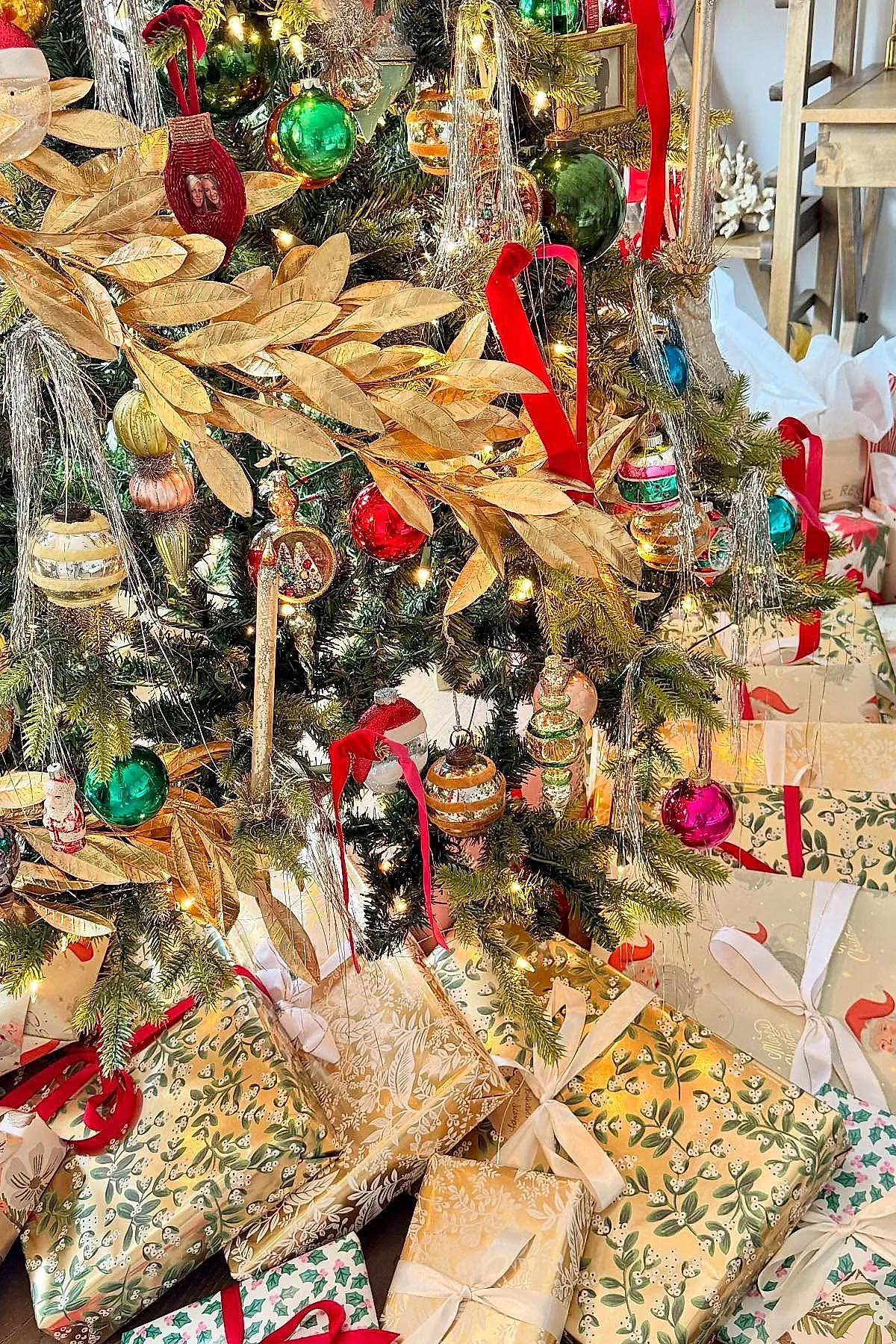 Vintage Christmas tree decorated with shiny green, red, pink, and gold glass ornaments, accented with silver tinsel and golden leaf branches, surrounded by beautifully wrapped presents in patterned gold and cream paper tied with ribbons.