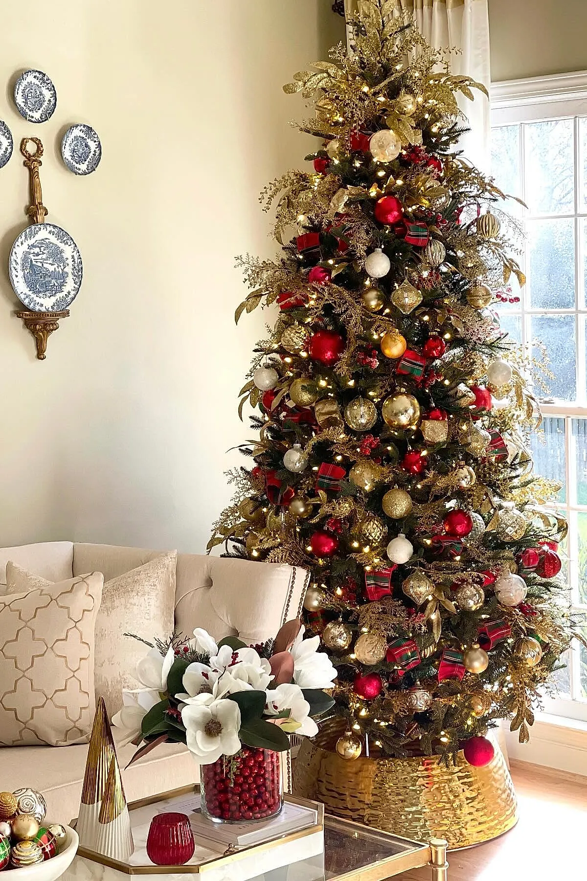 traditional christmas tree decorated with gold foliage sprigs, red and gold baubles, plaid ribbon bows, and warm white string lights, standing next to a window in a living room.
