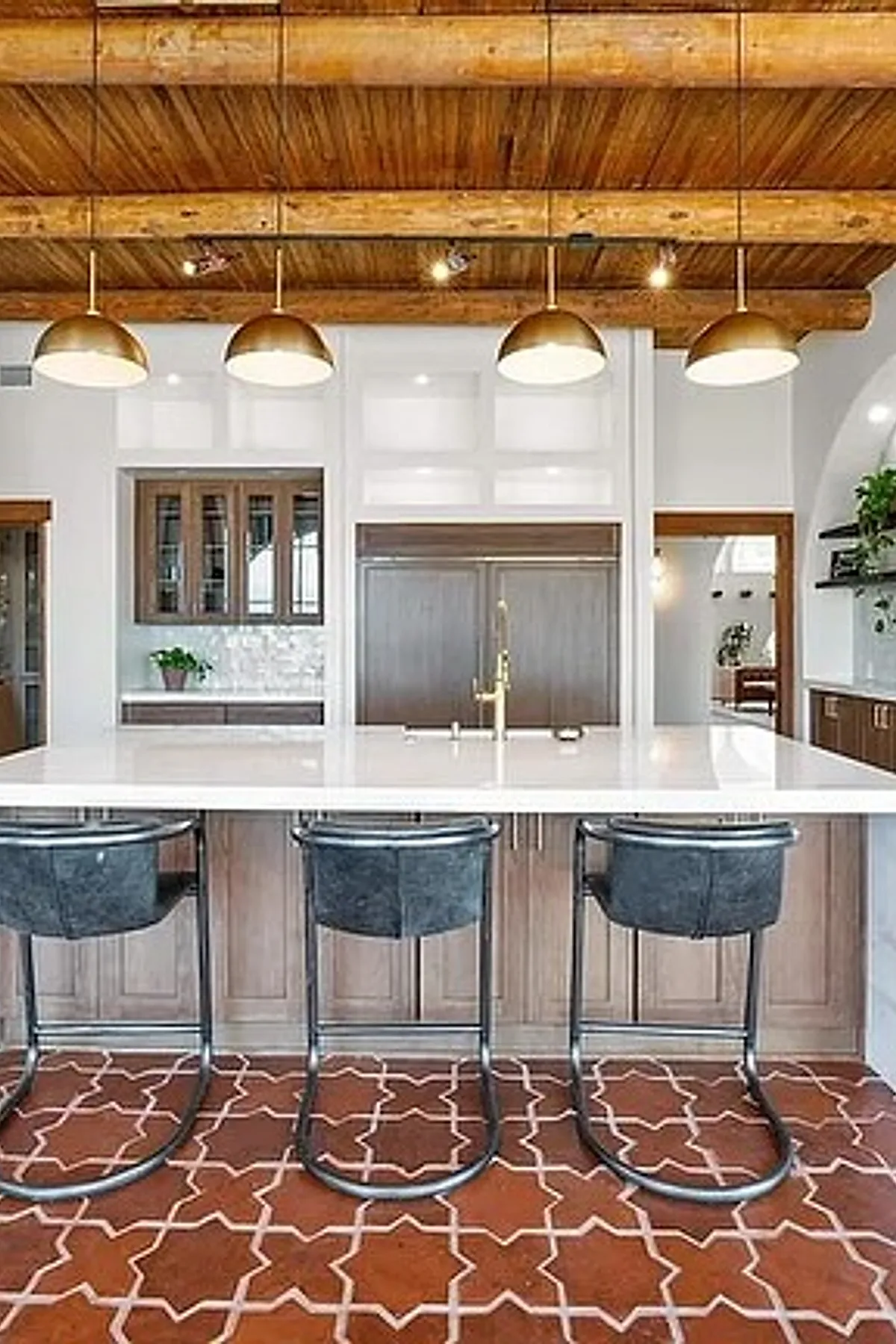 Modern Spanish style kitchen featuring exposed wooden ceiling beams, four gold dome pendant lights over a white marble island with wood cabinetry, black leather bar stools, and a terracotta tile floor with white geometric patterns.