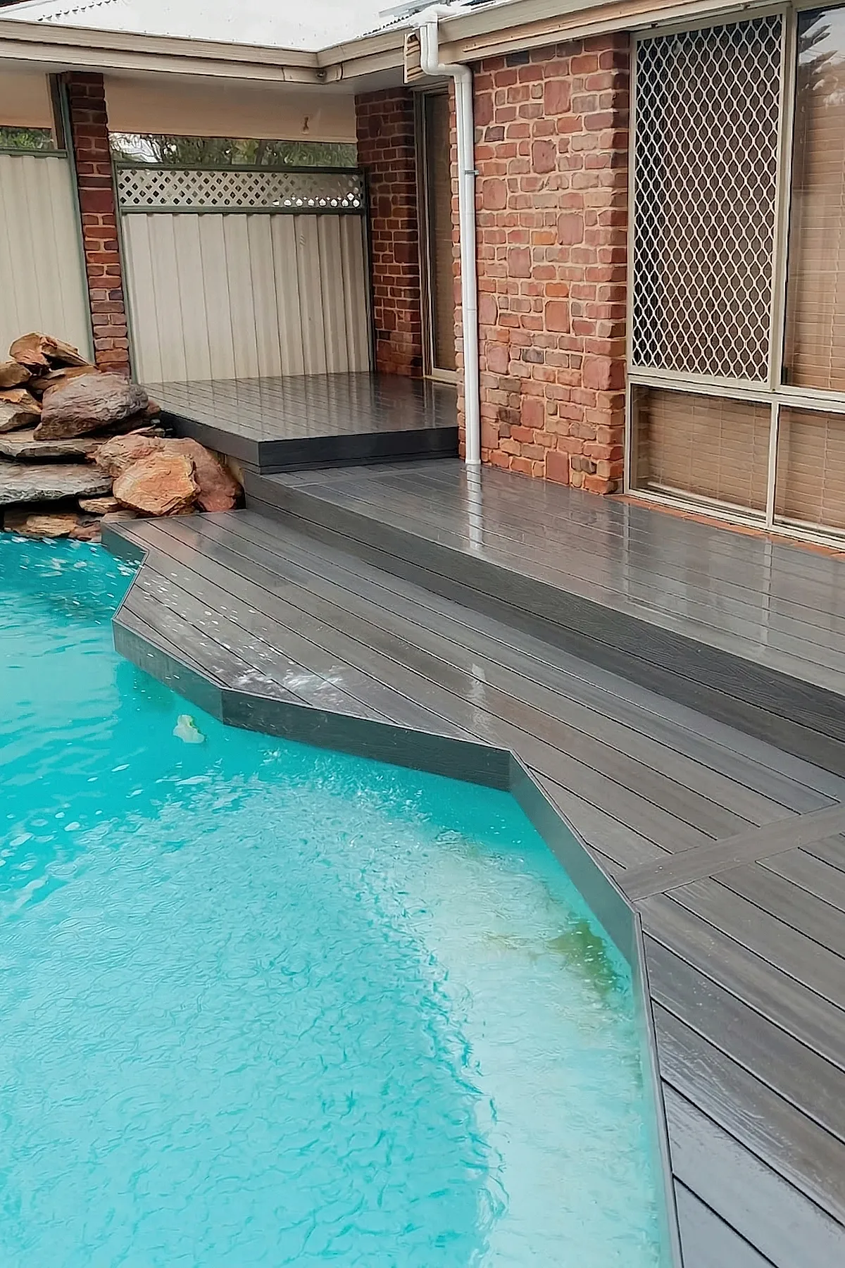 A glossy gray composite pool deck with clean lines surrounding a blue swimming pool, adjacent to a brick house wall and a stacked natural rock water feature.