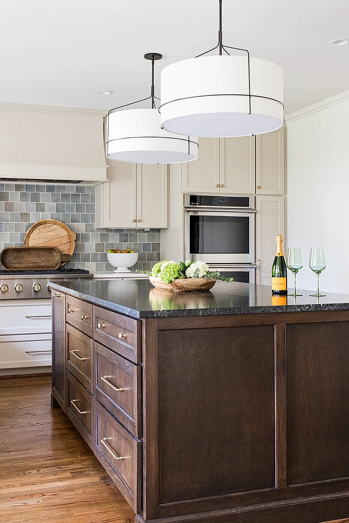 Cottage kitchen featuring a gray square tile backsplash in varying shades behind a stove, cream cabinetry with gold hardware, dark wood island with black countertop, two large white drum pendant lights, hardwood flooring, and decorative wooden trays on counter near stove.