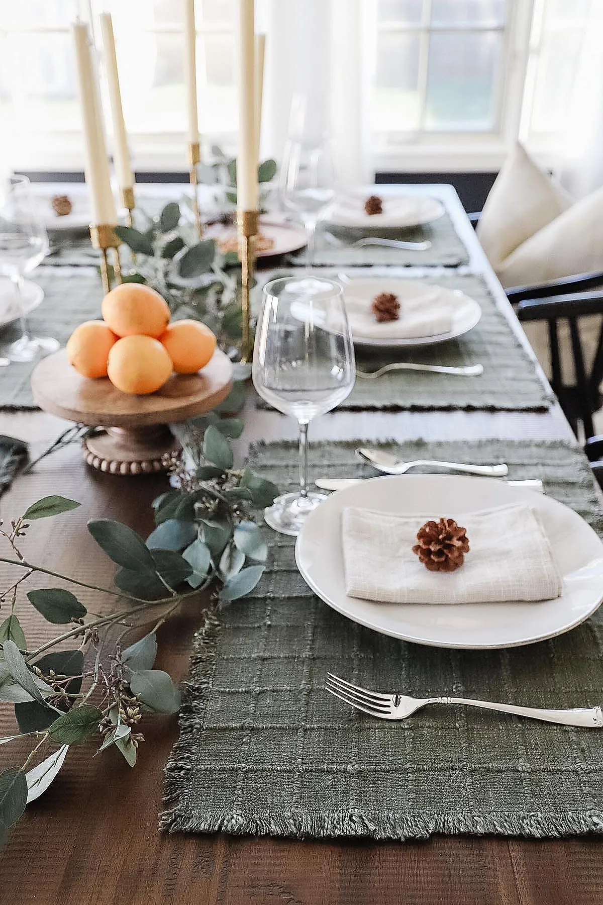 Thanksgiving table with green woven placemats, white plates topped with beige linen napkins and pinecones, clear wine glasses, silver cutlery, eucalyptus garland running along wooden table, cluster of oranges on wooden pedestal, gold taper candle holders with cream candles