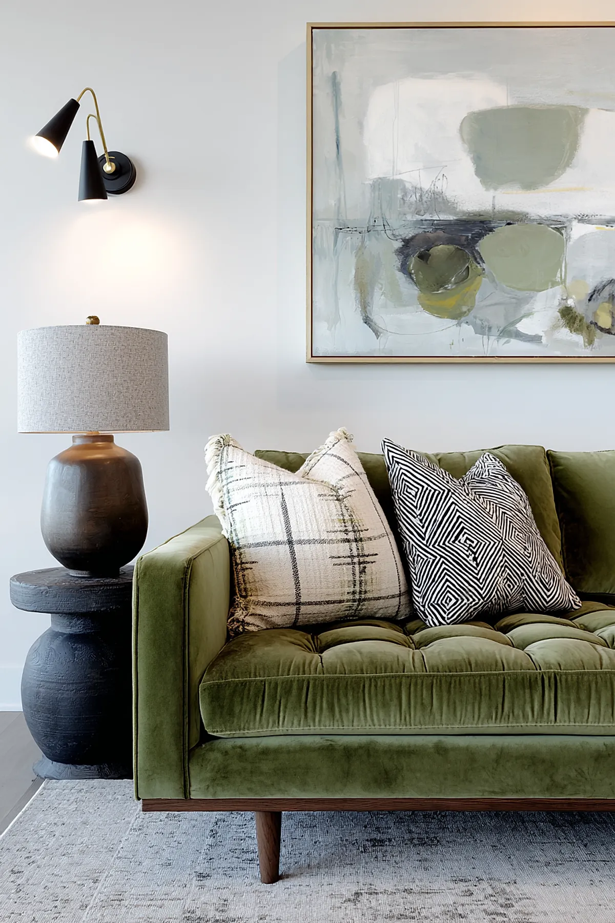 Earthy living room featuring a lush green velvet tufted sofa adorned with two large white plaid textured pillows and two black and white geometric patterned pillows, positioned against a light wall with abstract green and gray artwork above. A black double-cone wall sconce and dark round side table with a brown ceramic lamp complete the setup.
