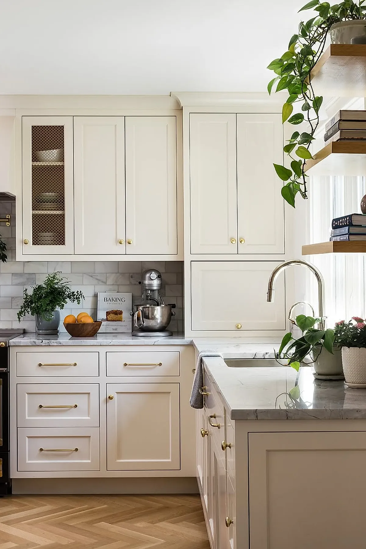 Cream-colored kitchen cabinets with brass hardware, white marble countertops, decorated with a wooden bowl of oranges, a potted plant, a stand mixer, and a baking book against white subway tile backsplash.