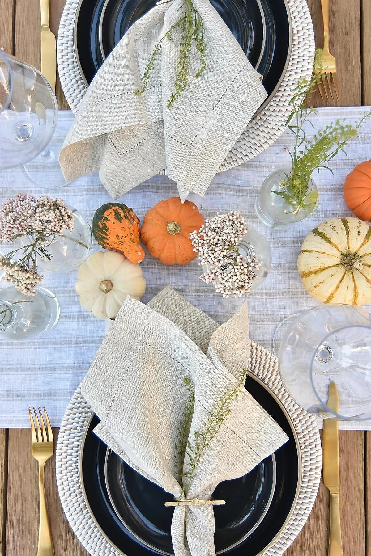 Fall dining table setting featuring beige linen napkins tied with green sprigs, black plates on white woven chargers, mini orange and white pumpkins, gold flatware, and clear glass vases with small flowers.
