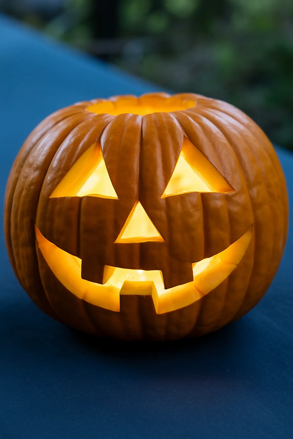 Orange pumpkin carved with two triangle eyes, triangle nose, and wide smiling mouth with two teeth, glowing from inside on blue surface outdoors.