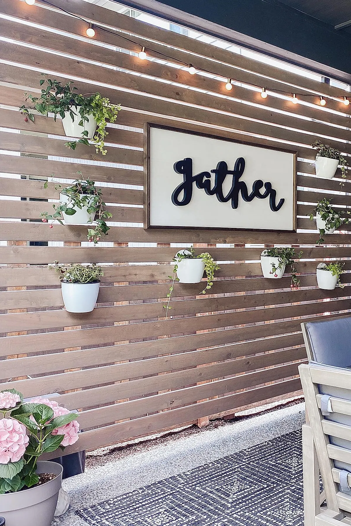 Wooden planter privacy wall with horizontal slats featuring white hanging pots with trailing green plants and string lights above in an outdoor patio setting