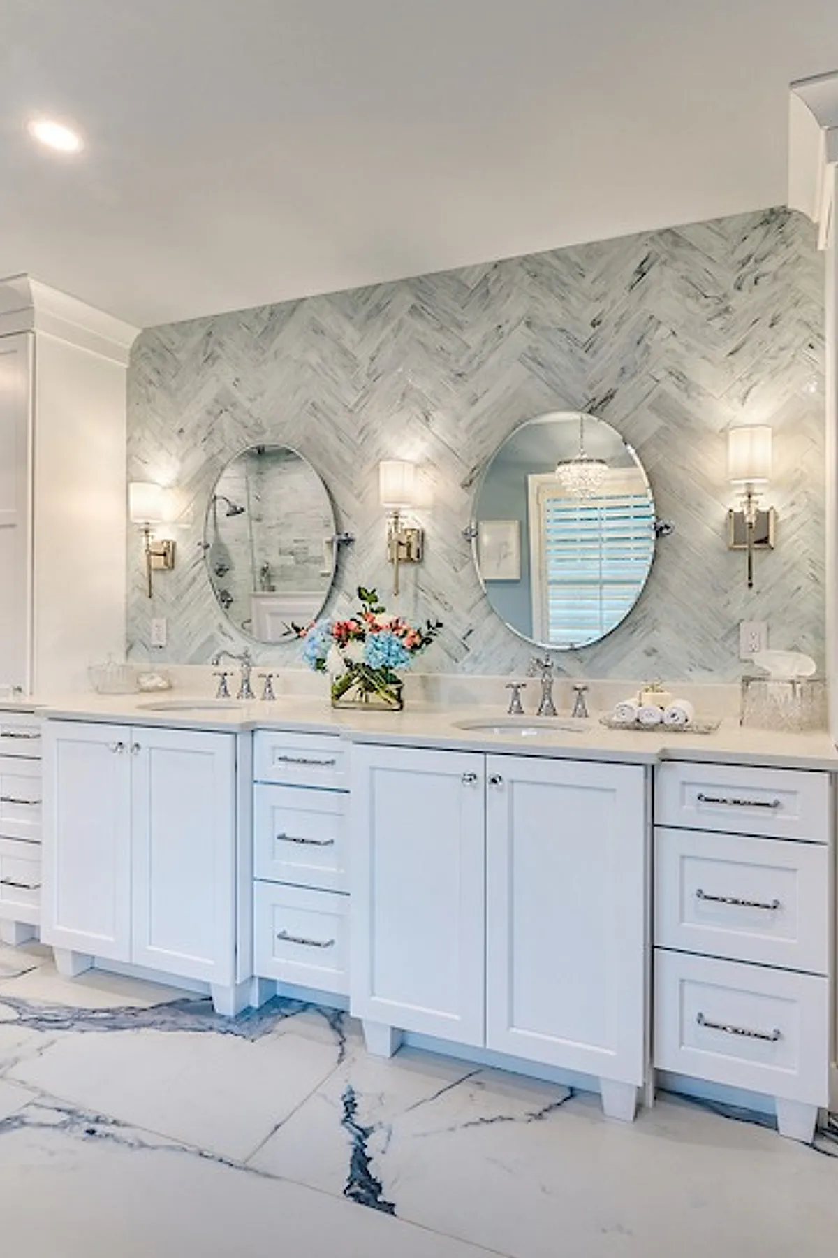 Luxury master bathroom with white cabinetry and drawers, marble countertop with two sinks, oval mirrors above each sink, herringbone pattern marble tile backsplash wall, two wall sconces with white shades flanking the mirrors, and large marble floor tiles with gray veining.