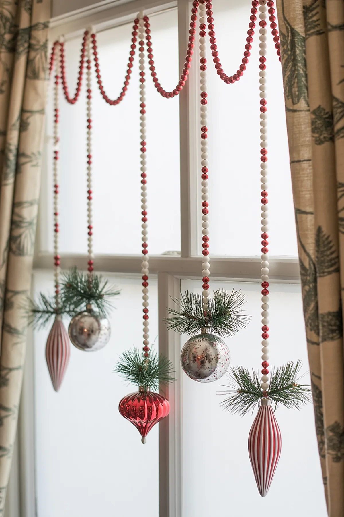 christmas window decorations featuring red and white beaded garlands draped in loops and strands, with hanging red and silver glass ornaments accented by green pine sprigs against a white window frame
