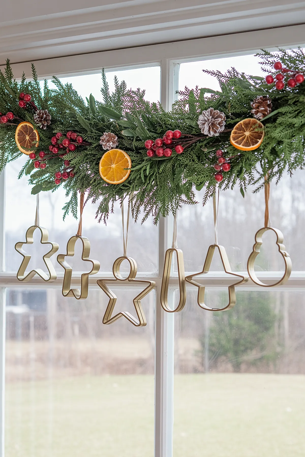 christmas window decorations featuring a lush green garland decorated with frosted pine cones, red berries, and dried orange slices, with metallic gold cookie cutter ornaments in festive shapes hanging from the garland against a white window frame.