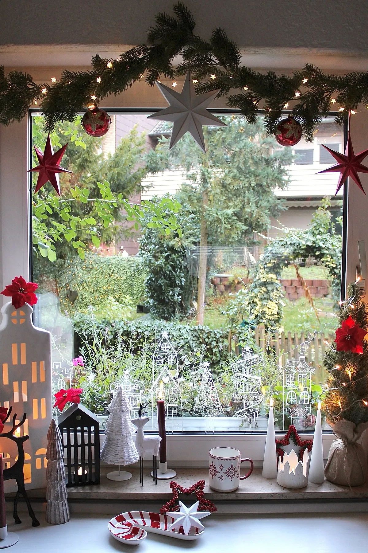 christmas window decorations featuring white miniature village houses, red candles, star-shaped hanging ornaments, evergreen garland with tiny star lights, and poinsettia flowers on the window sill and frame