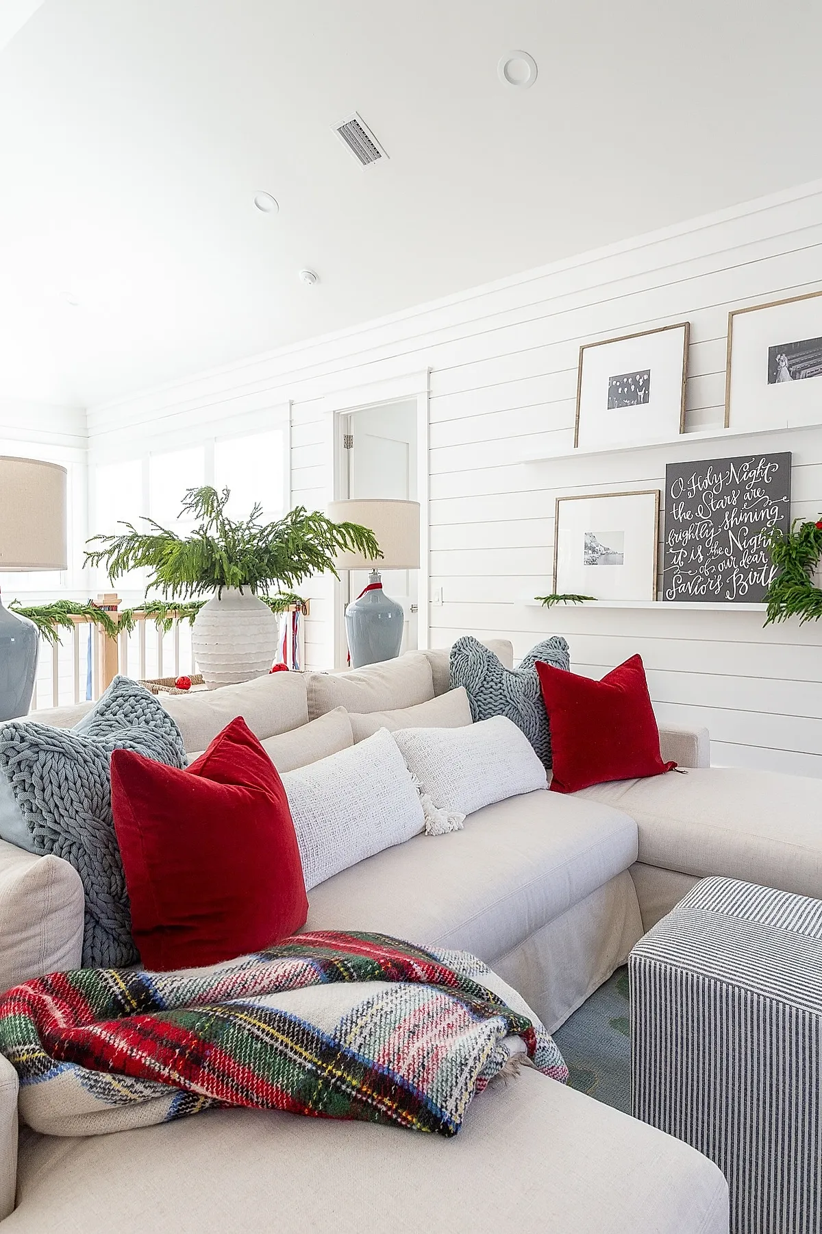 bright living room with minimalist christmas decor featuring a beige sectional sofa with red velvet pillows, chunky knit grey cushions, white lumbar pillows, and a colorful plaid blanket draped over the armrest, alongside green garland and framed art on white shiplap walls.