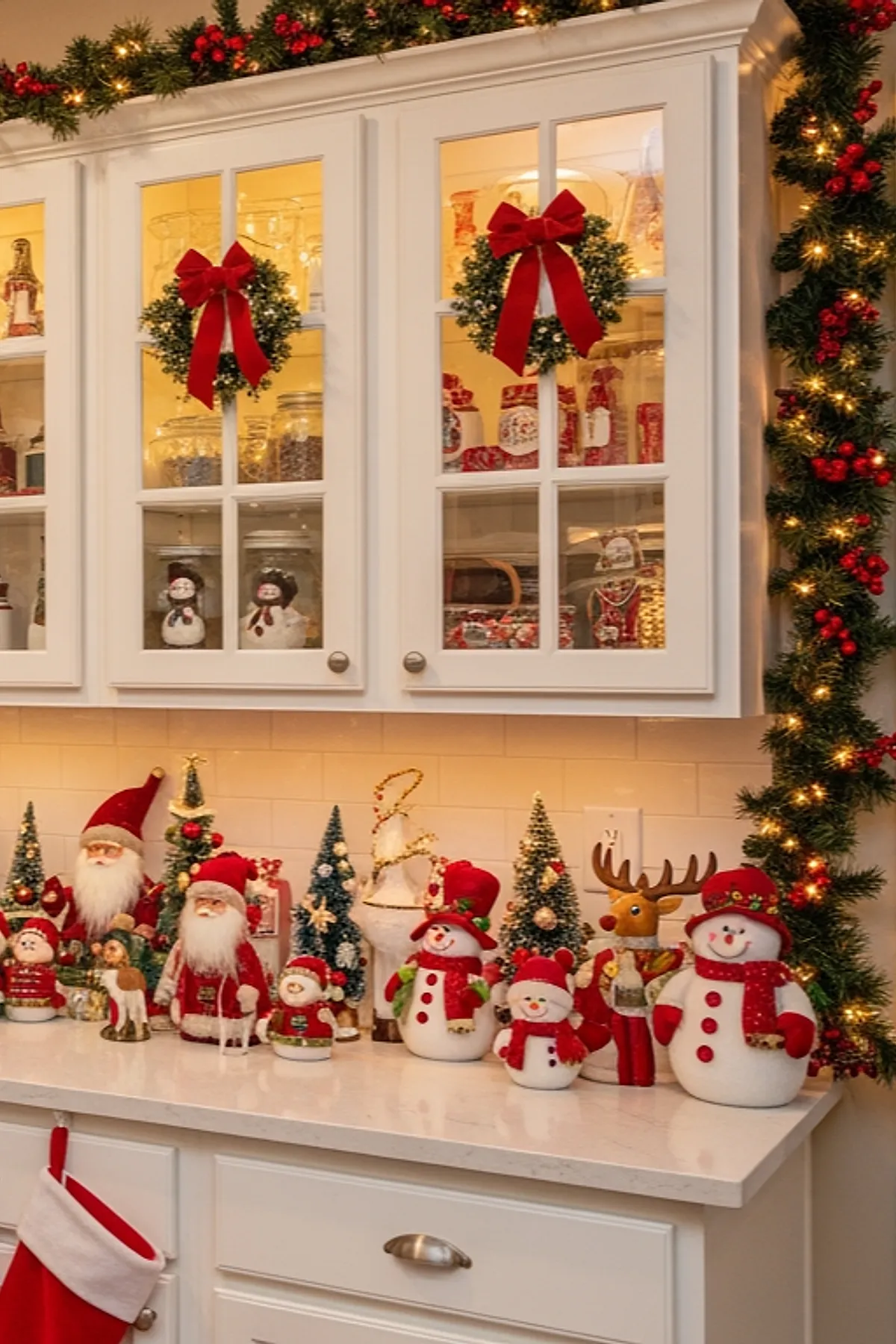 White countertop filled with Christmas figurines including Santas, snowmen, and reindeer surrounded by garlands and lights across white cabinets with glass doors.
