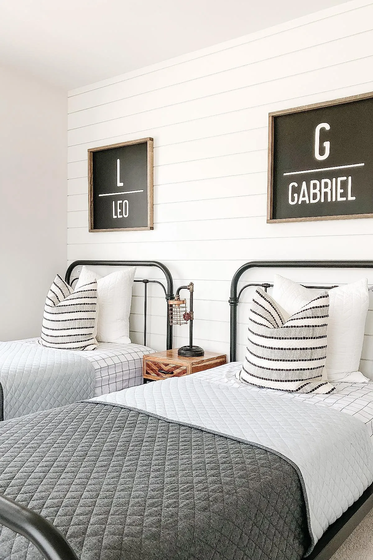 Two twin black metal frame beds with white quilted bedding and black-and-white striped pillows. Each bed has a framed blackboard-style wall art above it featuring a large initial and name: